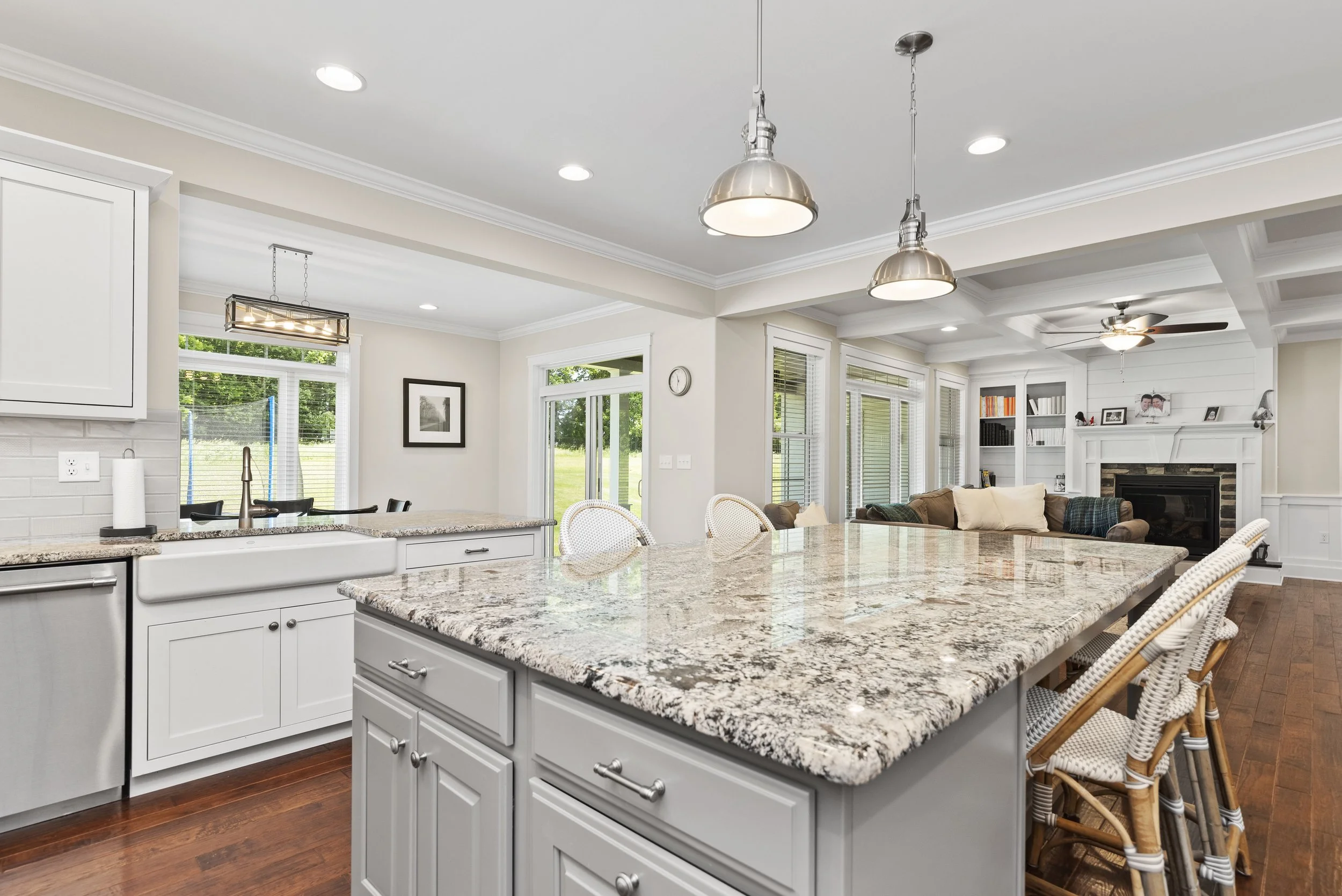 Open-plan kitchen and living room with granite countertops, white cabinetry, pendant lighting, woven bar stools, hardwood floors, beige walls, large windows, and a fireplace with built-in shelving.