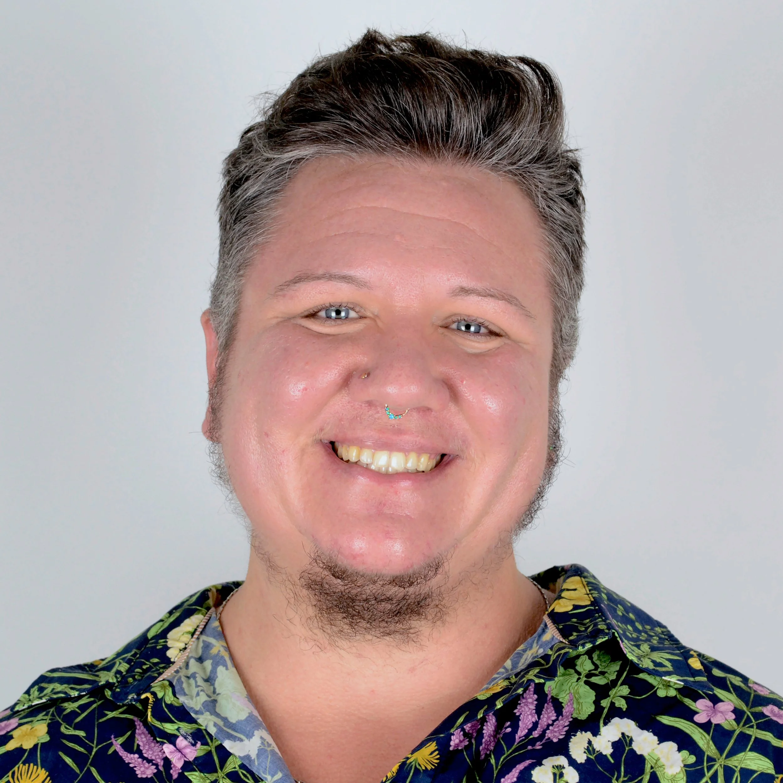 Heart, smiling, wearing colorful floral button down shirt in front of silver background.