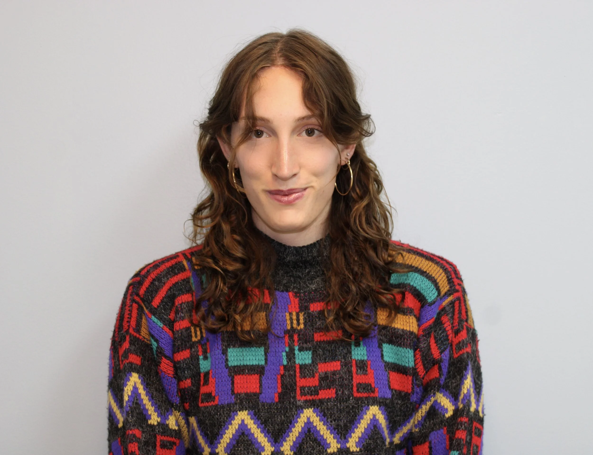 person with mid-length wavy brown hair looking at the camera, smiling warmly in a brightly-patterned sweater.
