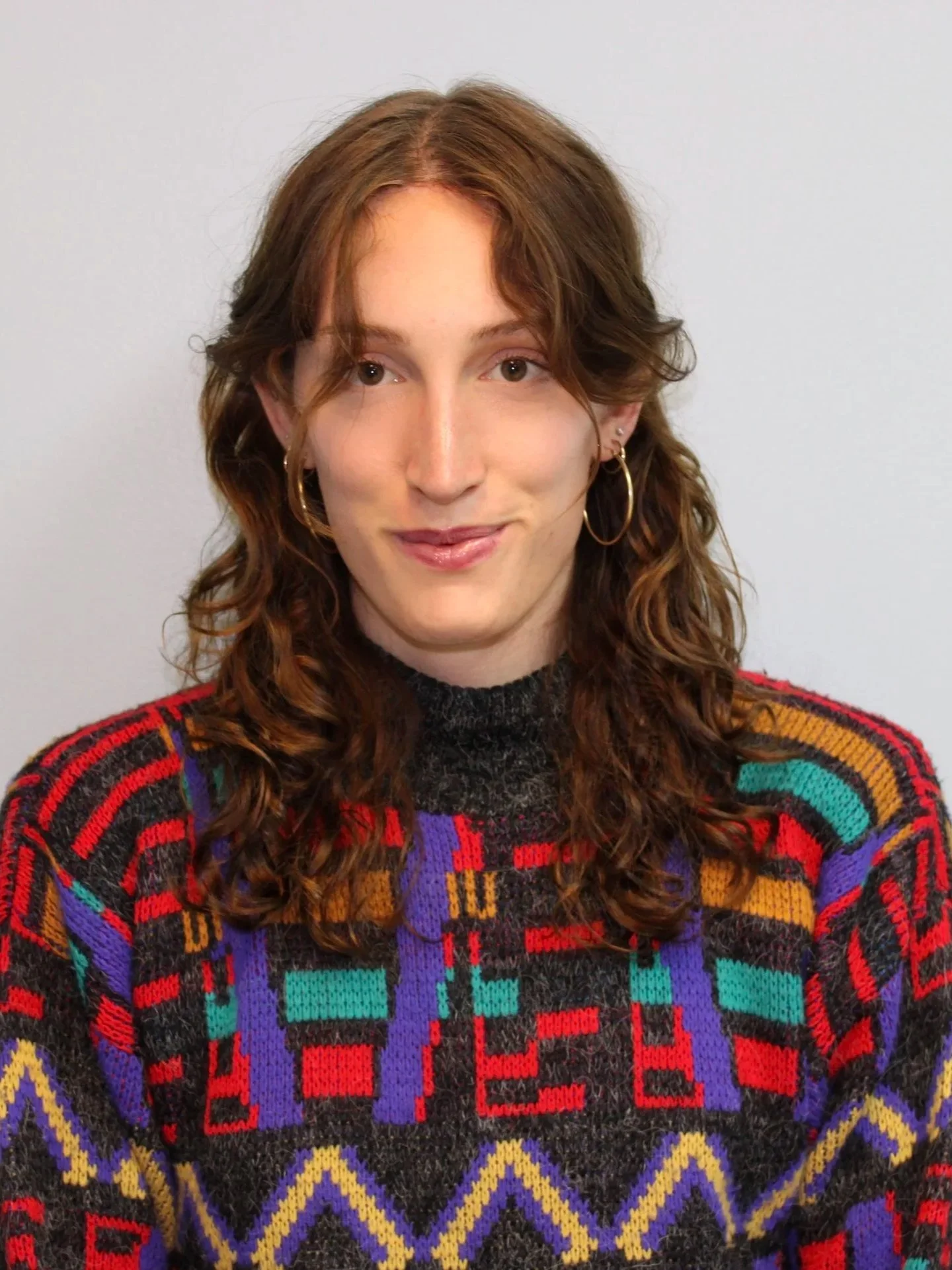person with mid-length wavy brown hair looking at the camera, smiling warmly in a brightly-patterned sweater.