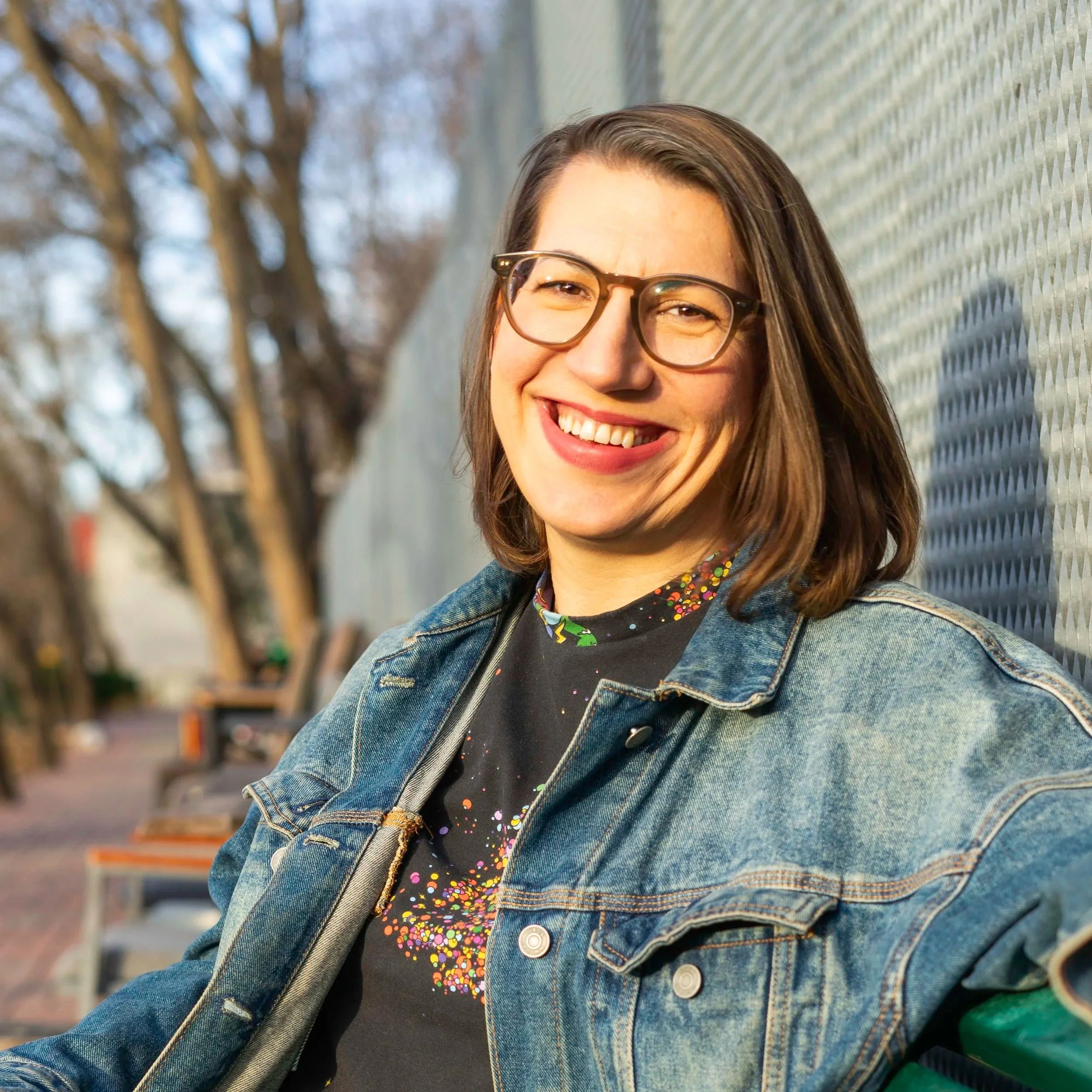 person with brown hair in a bob sitting outside on a park bench, smiling at the camera wearing a blue denim jacket, a black shirt with colorful splatters on it and black pants.