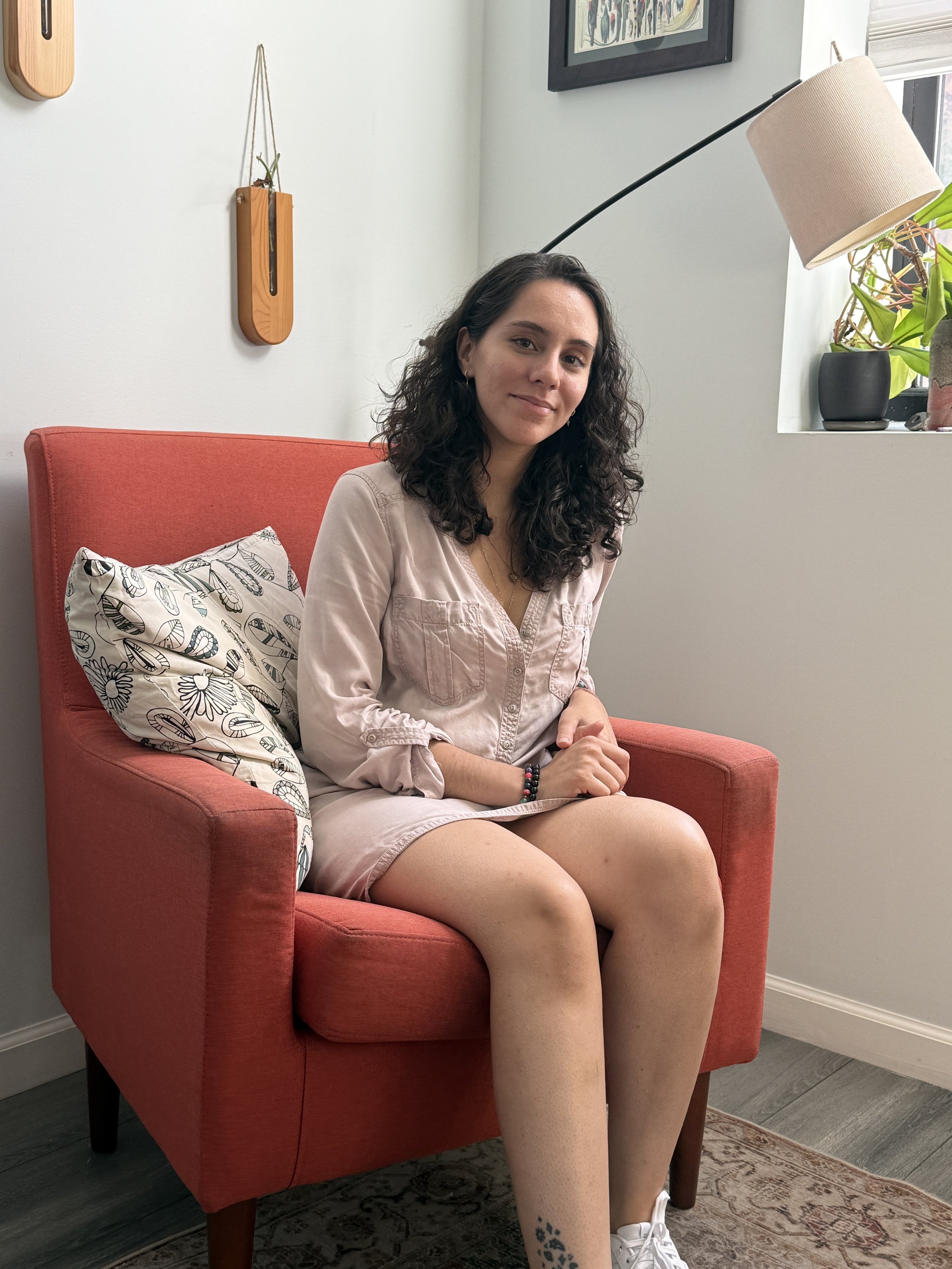 Image of a woman with medium length curly brown hair, seated in a red comfy chair. The woman is wearing a neutral colored dress and white sneakers.