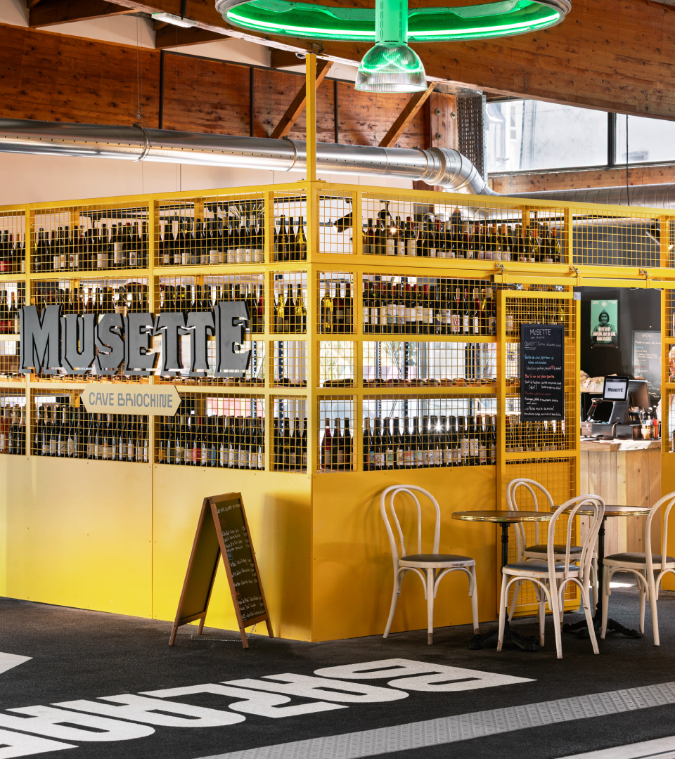 Un stand de dégustation de bière appelé 'Musette' avec une structure jaune contenant de nombreuses bouteilles de bière, situé dans un intérieur en bois avec un éclairage au plafond vert lumineux, des chaises blanches et un tableau noir avec le menu à côté.