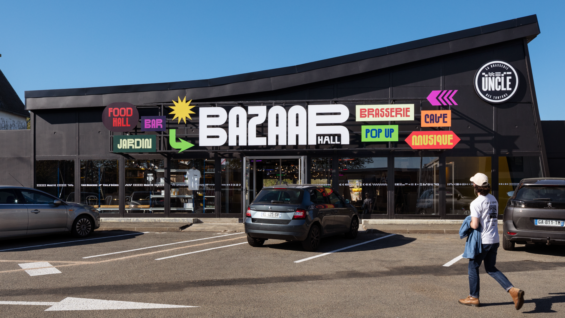 Façade d'un centre commercial avec un panneau coloré indiquant diverses zones et commerces, dont un food hall, bar, jardin, brasserie, cave, musique, et un logo « UNCLE » en haut à droite. Il y a des voitures stationnées devant et une personne marchant à proximité.