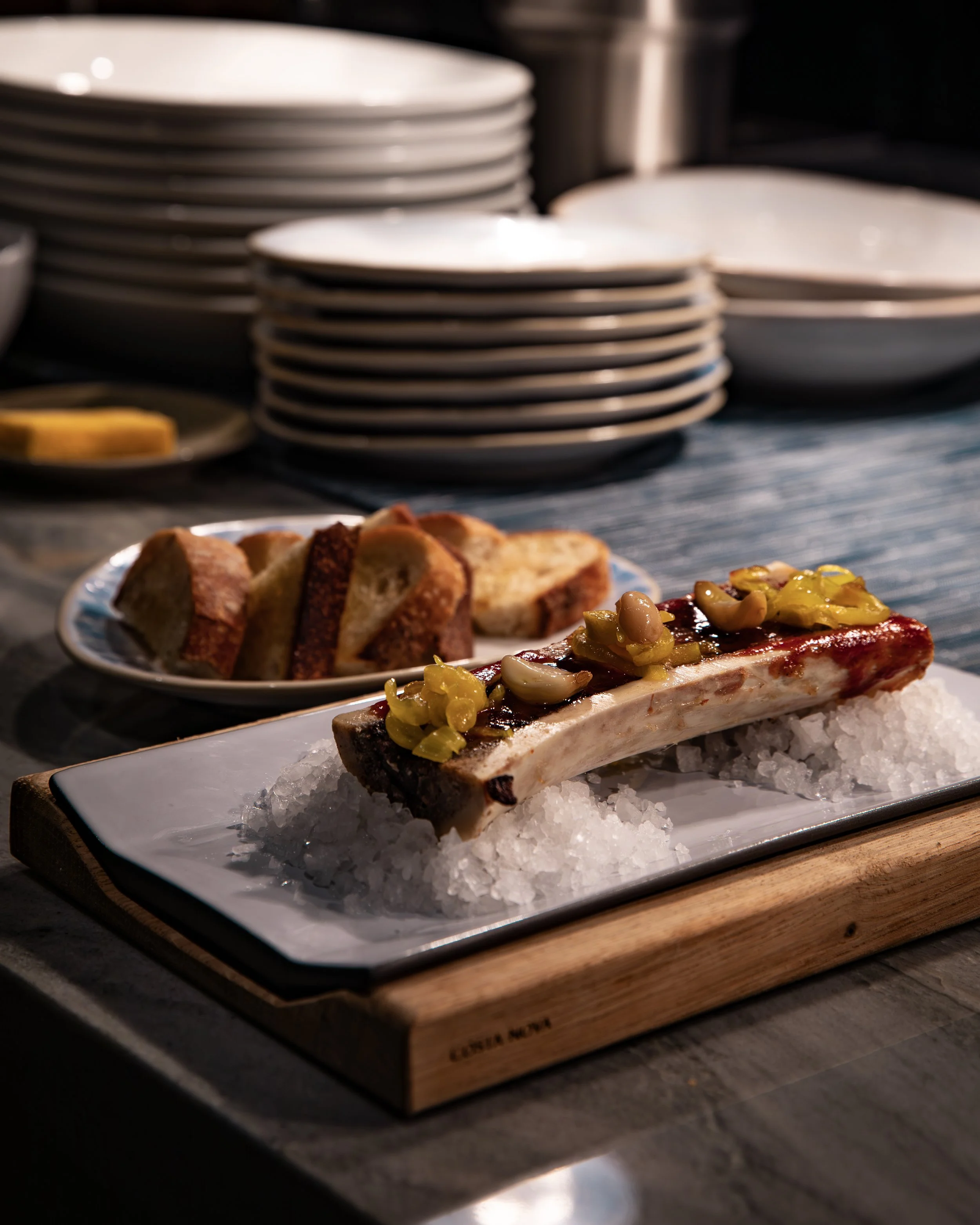 A plate of grilled beef ribs topped with chopped vegetables on a bed of ice, with sliced bread and stacked plates in the background.