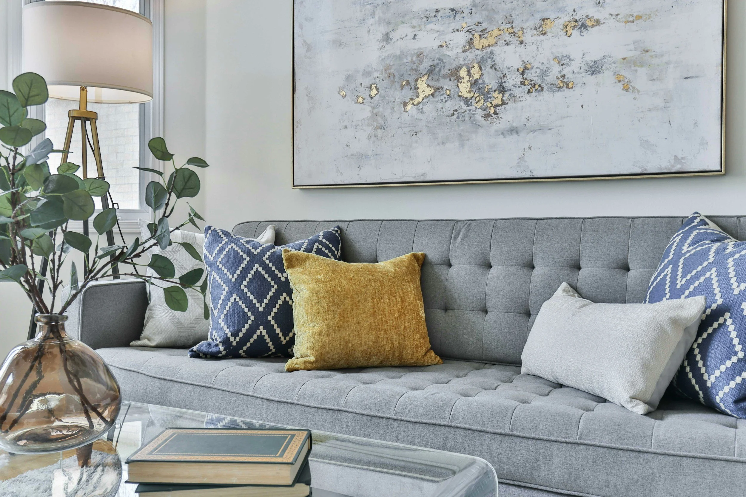 Living room with a gray tufted sofa decorated with colorful cushions including a yellow one and blue and white patterned ones. Behind the sofa, a large abstract painting with light and gold colors hangs on the wall. To the left, a window with a lamp and a potted plant on a table are visible, and a glass coffee table with books is in the foreground.