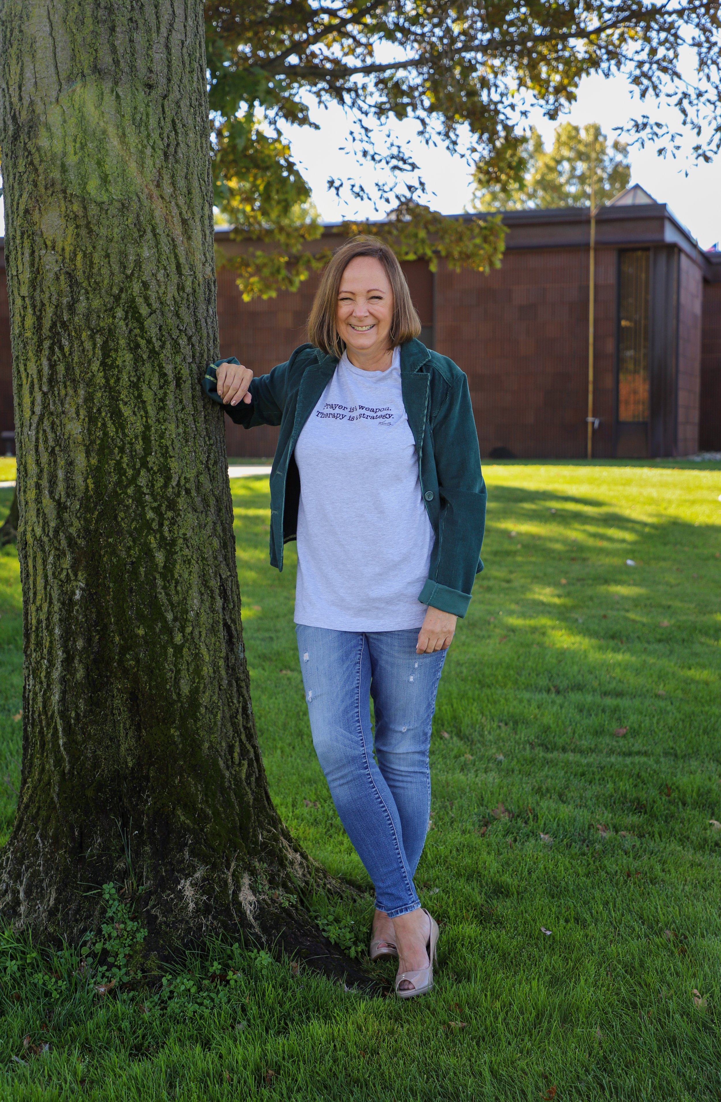 A woman with shoulder-length brown hair, wearing a white T-shirt with blue text, a green jacket, and distressed jeans, standing outdoors beside a tree, smiling at the camera. Behind her, there is a brick building and a grassy area under a partly cloudy sky.
