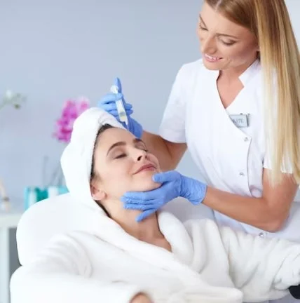 A beautician performs a facial treatment on a woman in a spa or clinic setting, using a syringe or needle while the woman relaxes with her eyes closed.