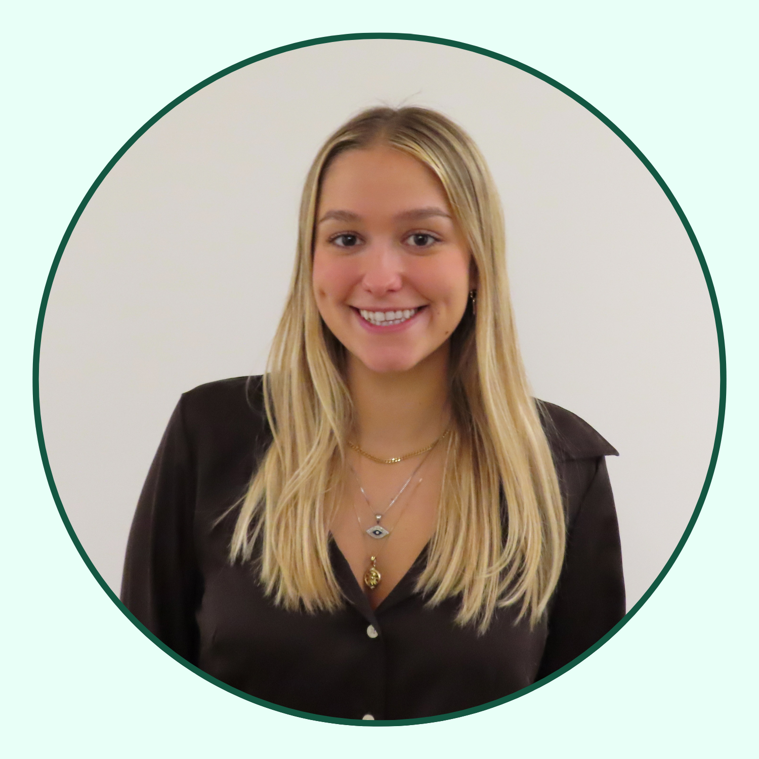 A young woman with blonde hair smiling, wearing a black button-up shirt and layered necklaces, standing against a plain white background.