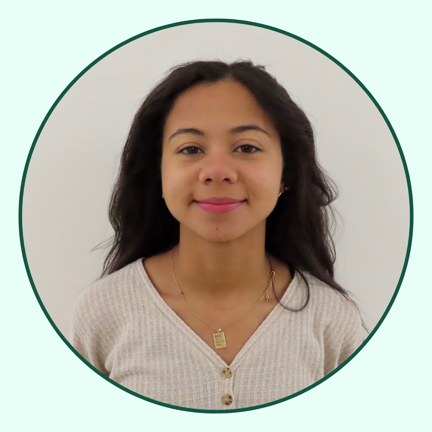 Young woman with shoulder-length wavy hair, wearing a beige sweater and a gold necklace, smiling with a neutral background.