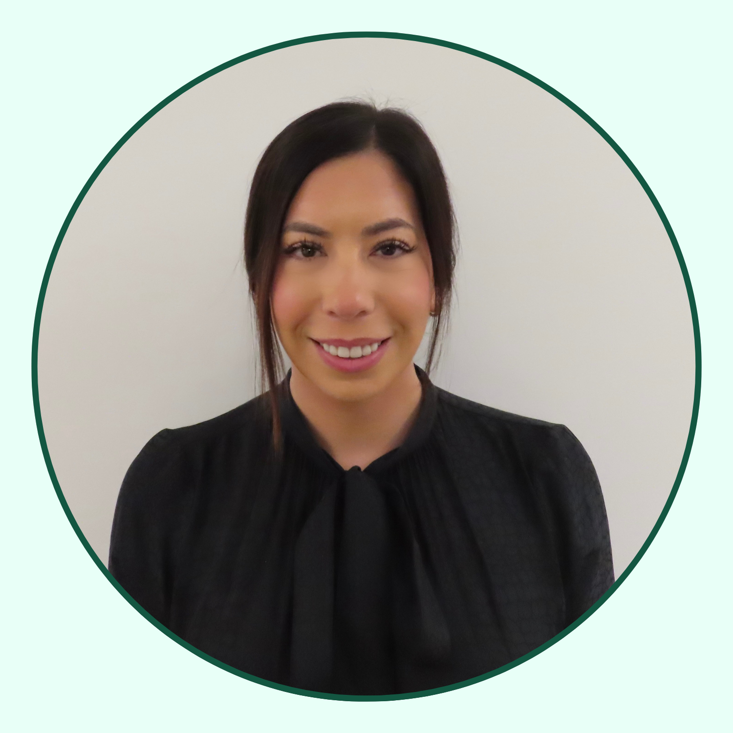 A woman with shoulder-length dark hair, smiling, wearing a black blouse with a bow at the collar, standing against a plain white background.