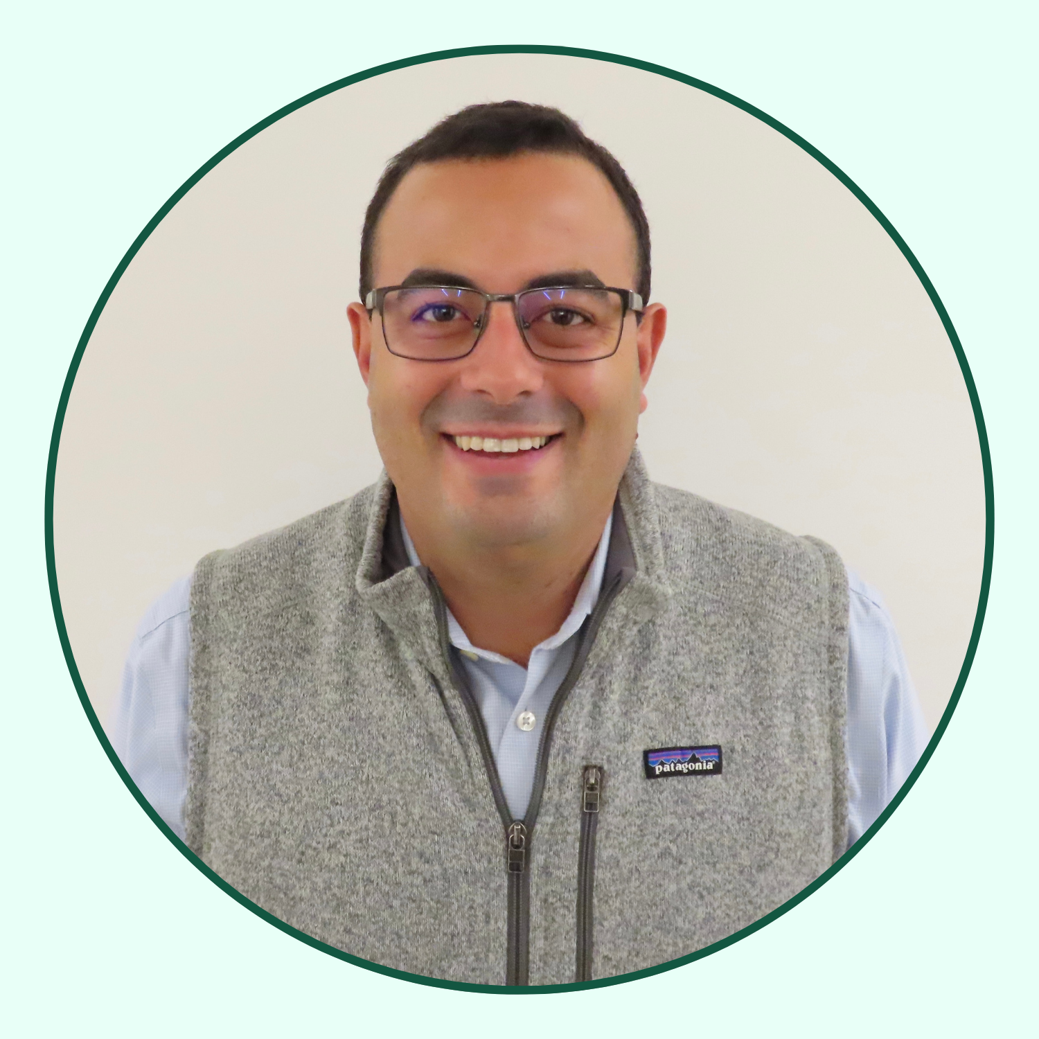 A smiling man wearing glasses, a white shirt, and a gray Patagonia fleece vest, looking at the camera, with a plain background.