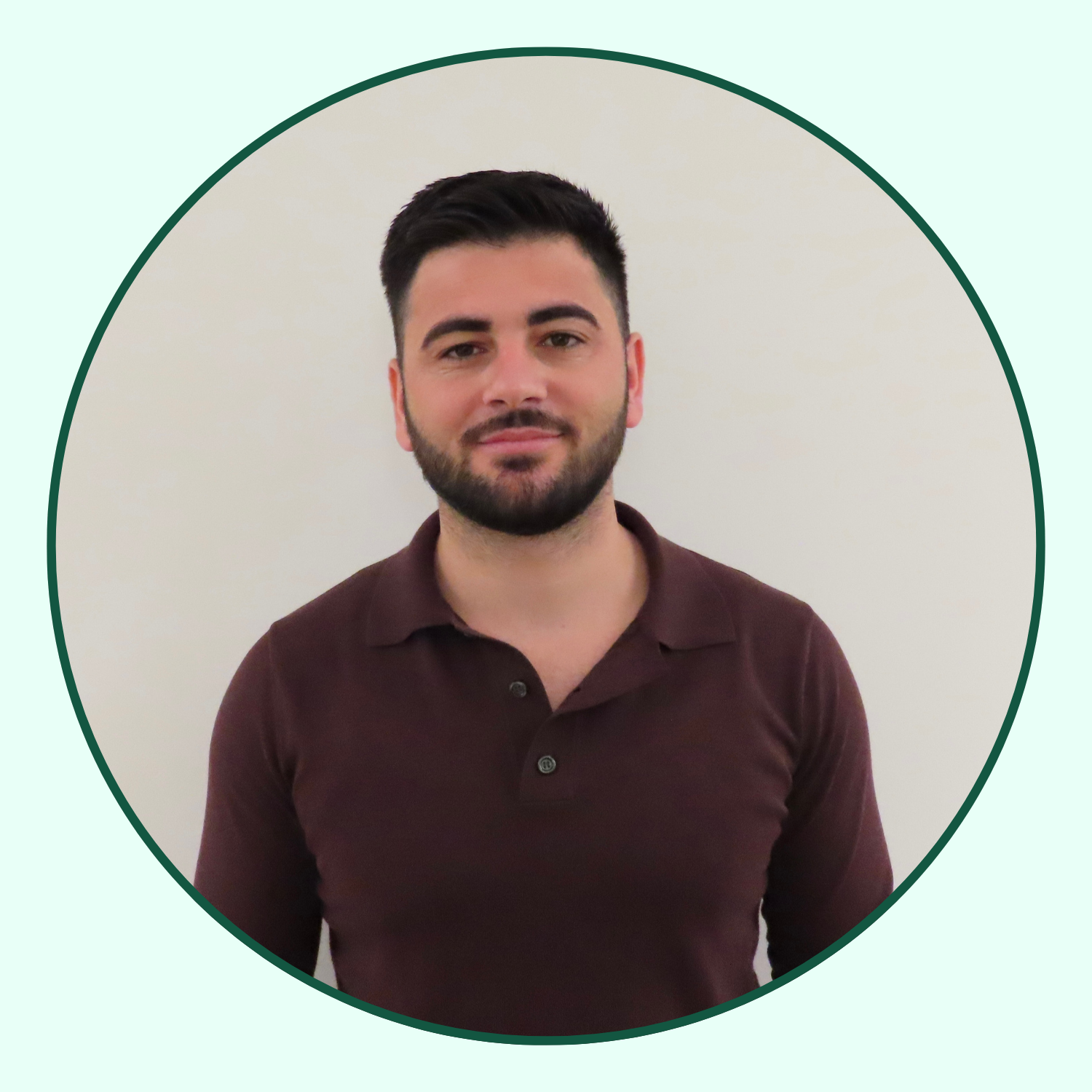 A young man with dark hair and a beard, wearing a maroon polo shirt, standing against a plain light-colored wall.