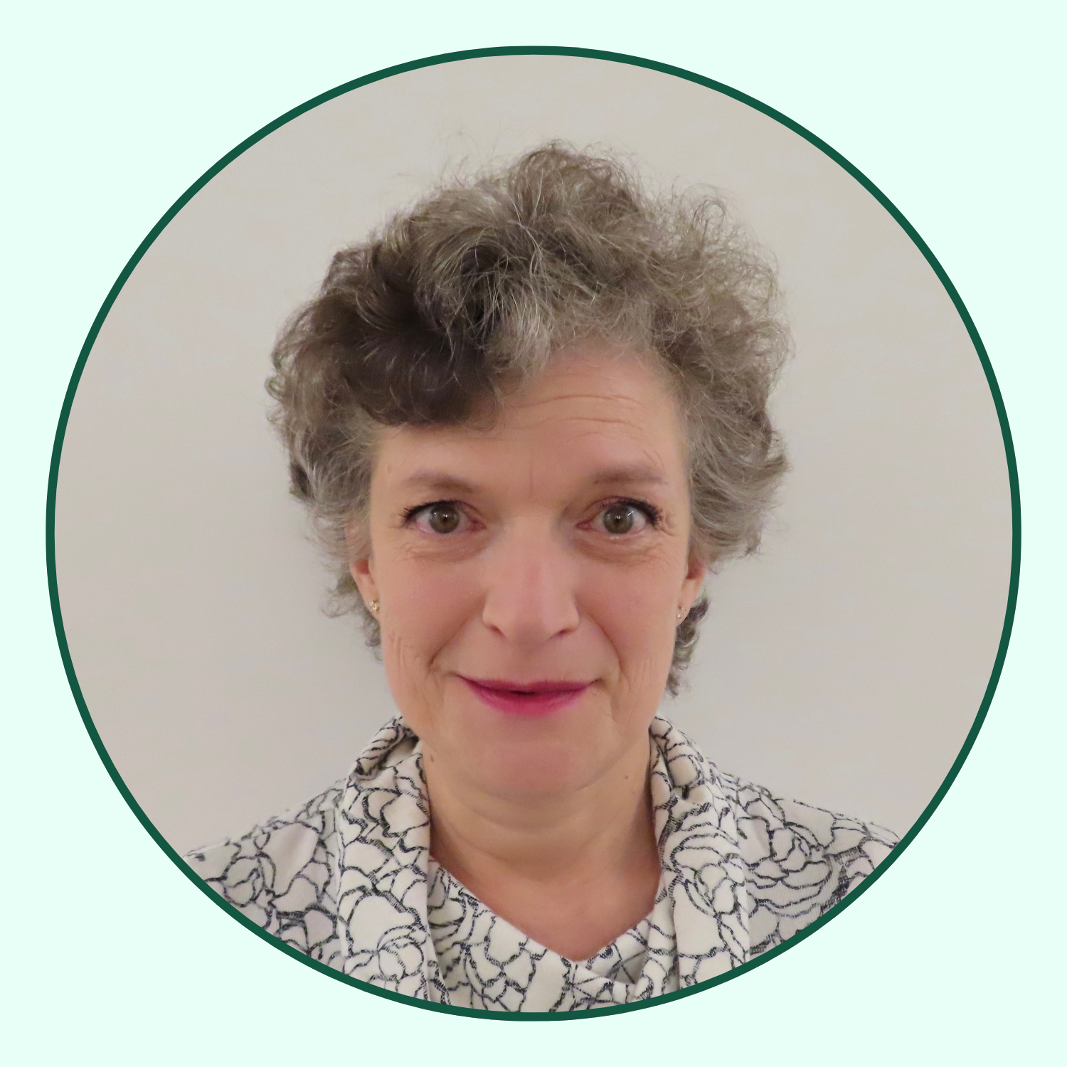 A portrait of a middle-aged woman with short, curly gray hair, wearing a white blouse with a black floral pattern, smiling against a plain light-colored background.