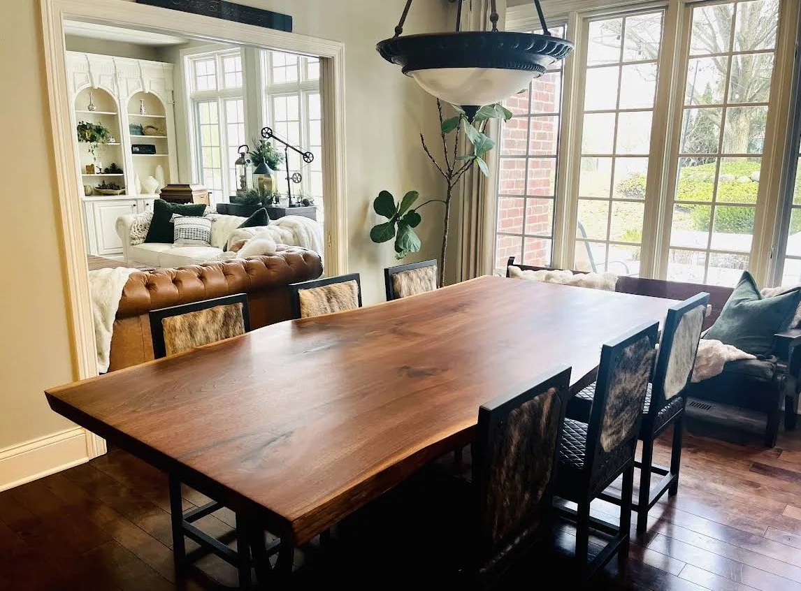 A dining room with a large wooden table surrounded by six chairs with animal print and black frames. A large hanging light fixture is above the table. There are large windows with a view of the outdoors. In the background, there is a living room with