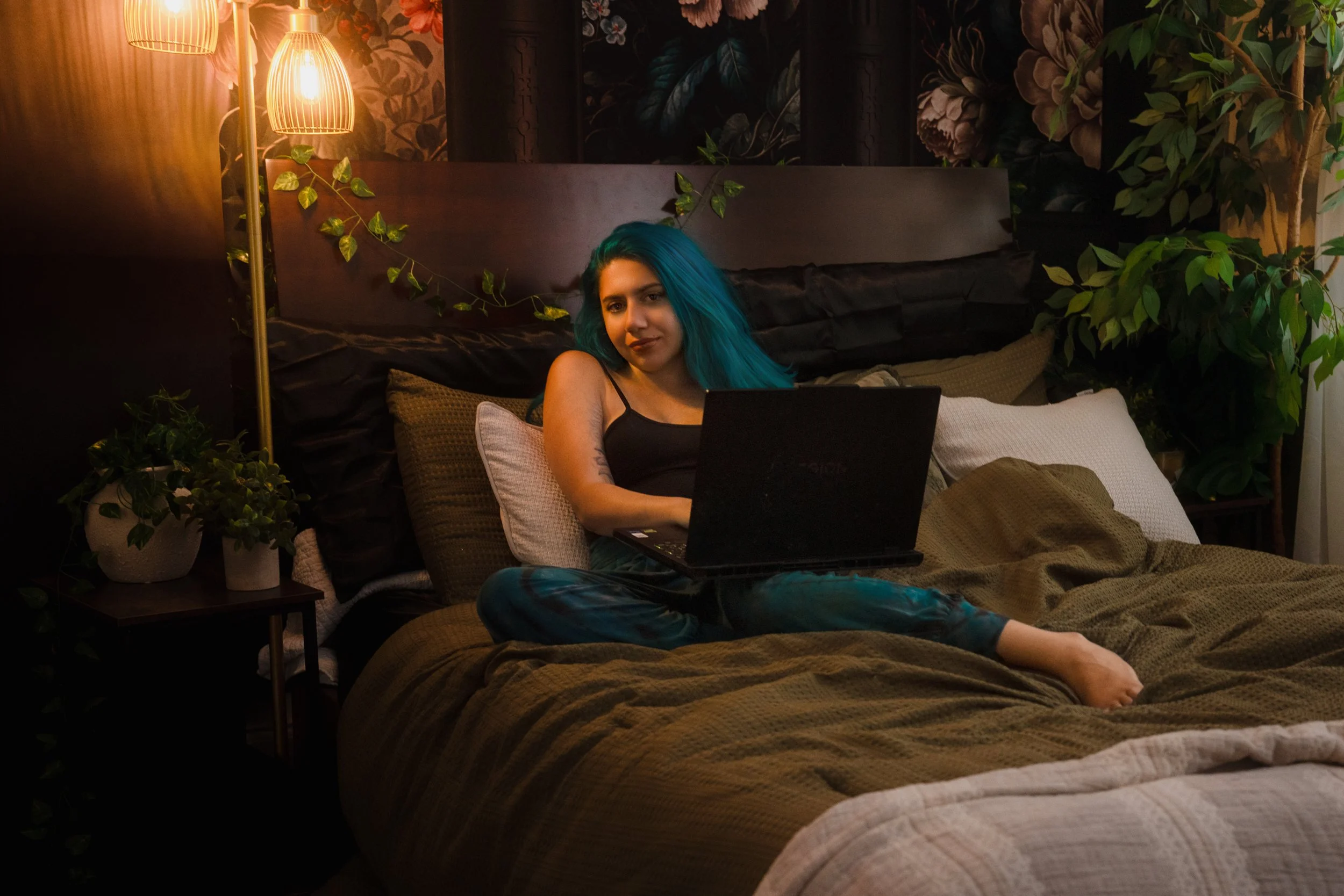 Young woman with blue hair sitting on a bed using a laptop in a dimly lit room decorated with plants and floral wall art.