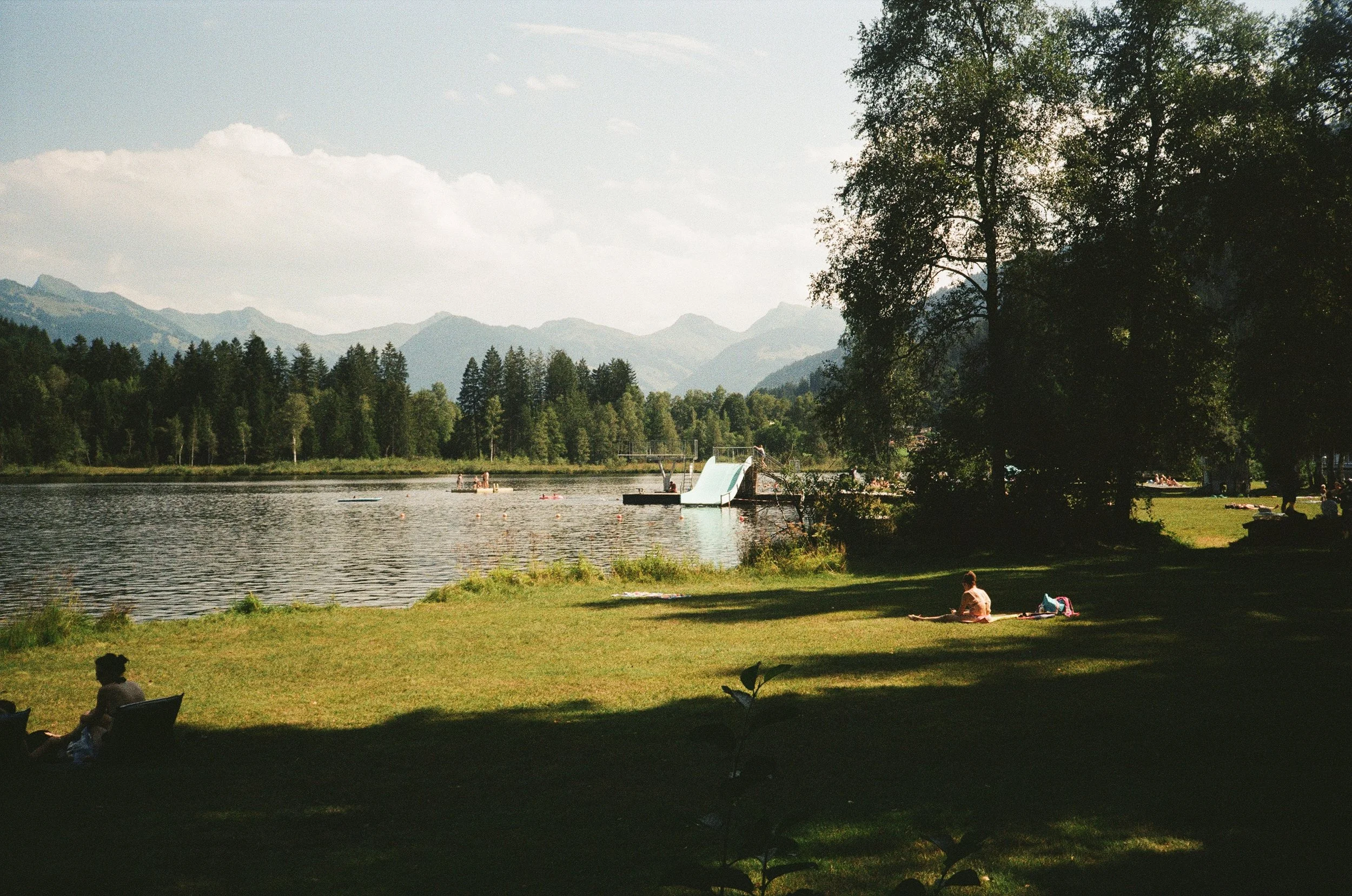 People relaxing on a grassy lakeside with trees, mountains, and a water slide in the background on a sunny day.