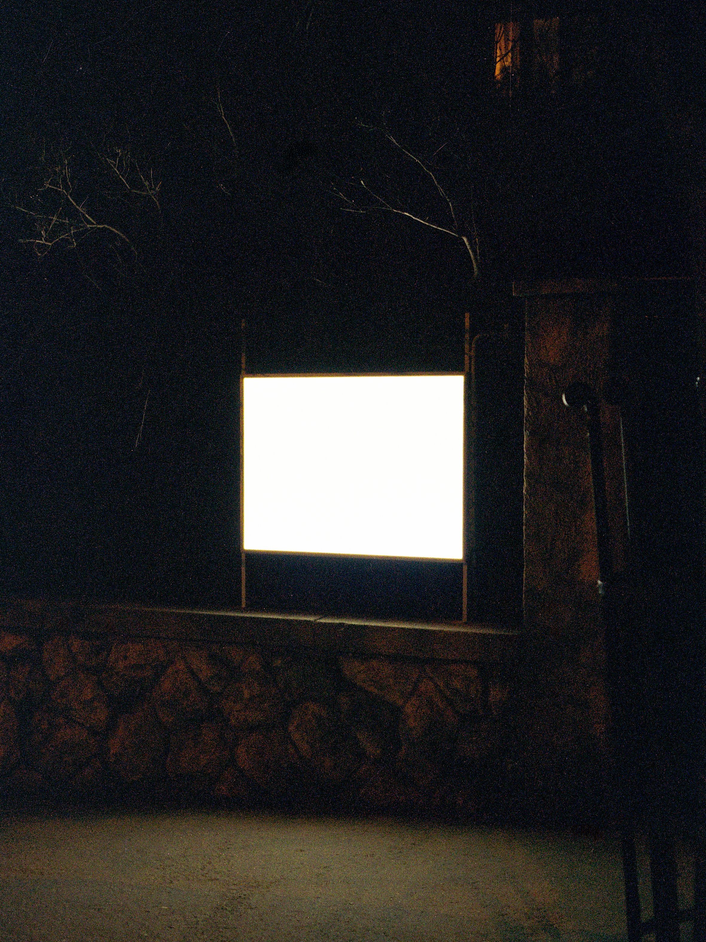 Dark outdoor scene at night with a brightly illuminated rectangular sign, stone wall, trees, and a building with warm lights in the background.