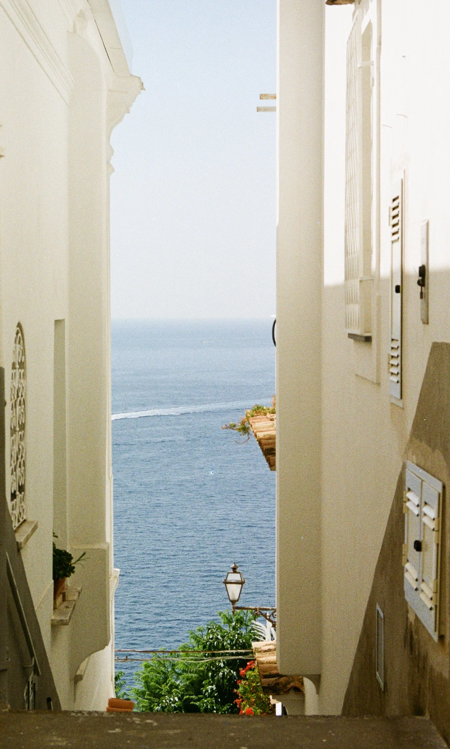 View through a narrow alleyway between white buildings overlooking the ocean, with some greenery, umbrellas, and a lamppost in the scene.