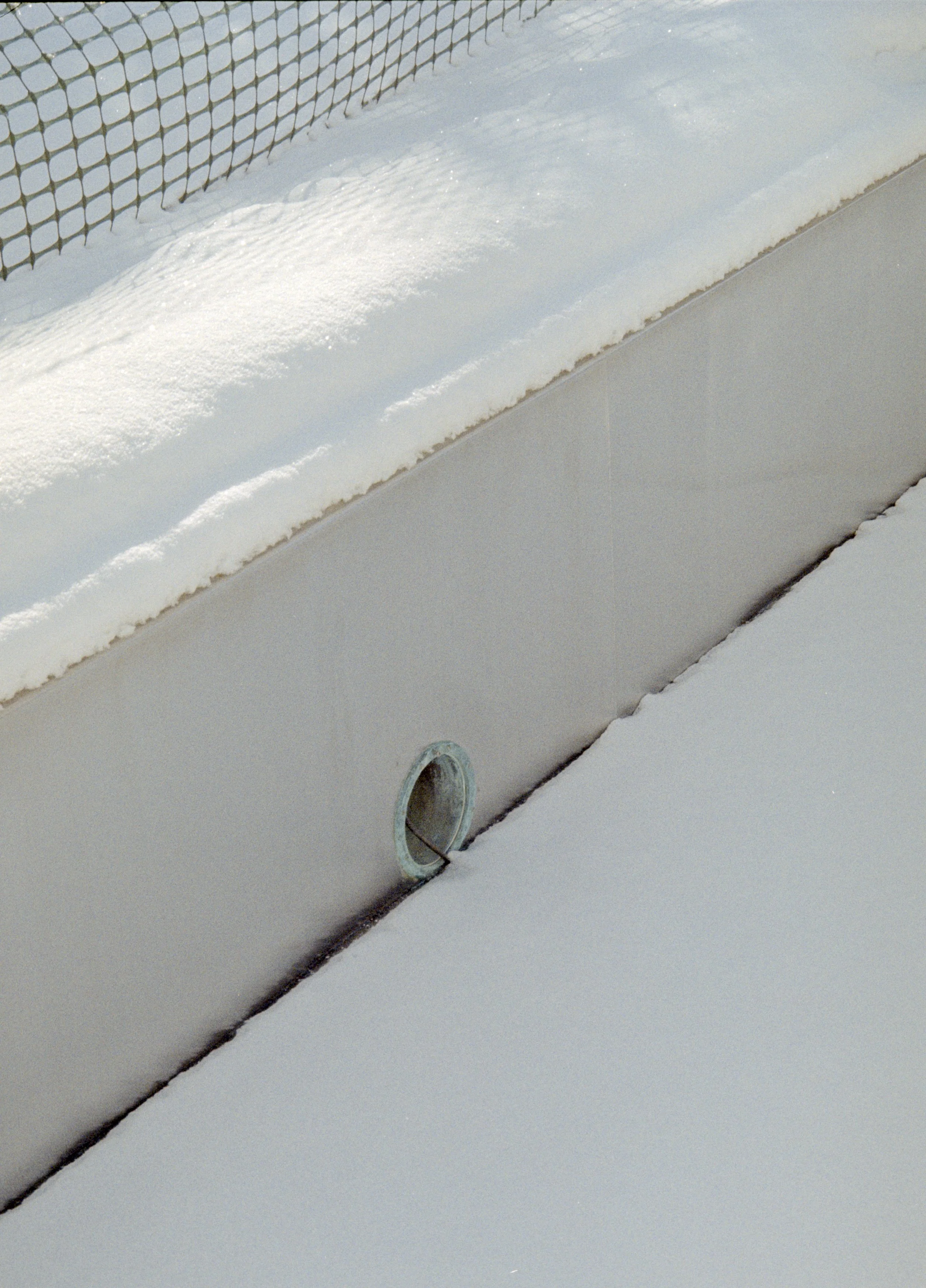 Close-up of a pool drain covered in snow, with a surrounding snow-covered area and a safety fence in the background.