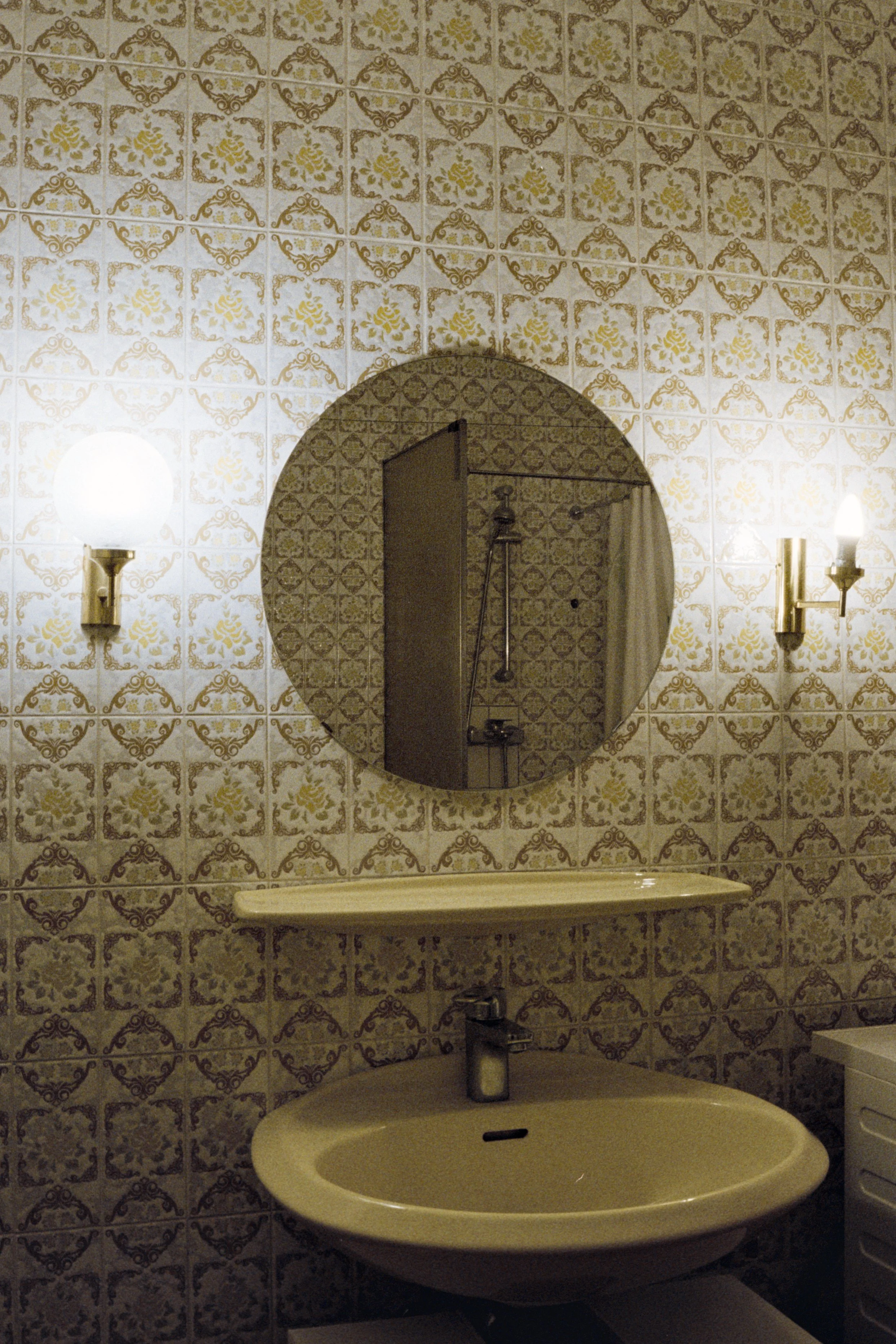 Bathroom with ornate patterned tiles, a round mirror, wall-mounted light fixtures, a sink, and a shower visible through the mirror reflection.
