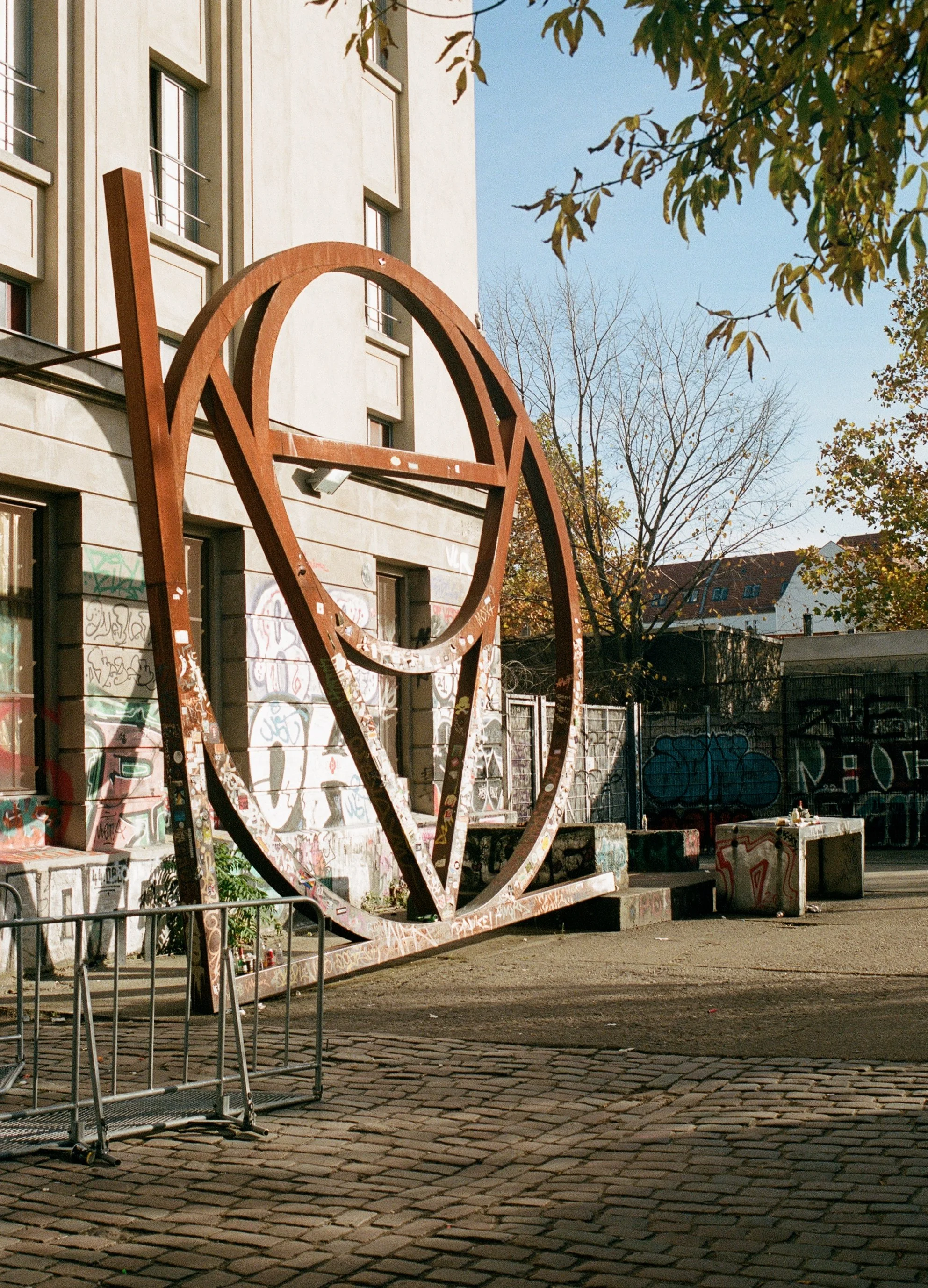 Large metal peace sign sculpture in an urban outdoor space with graffiti-covered walls, trees, and buildings in the background.