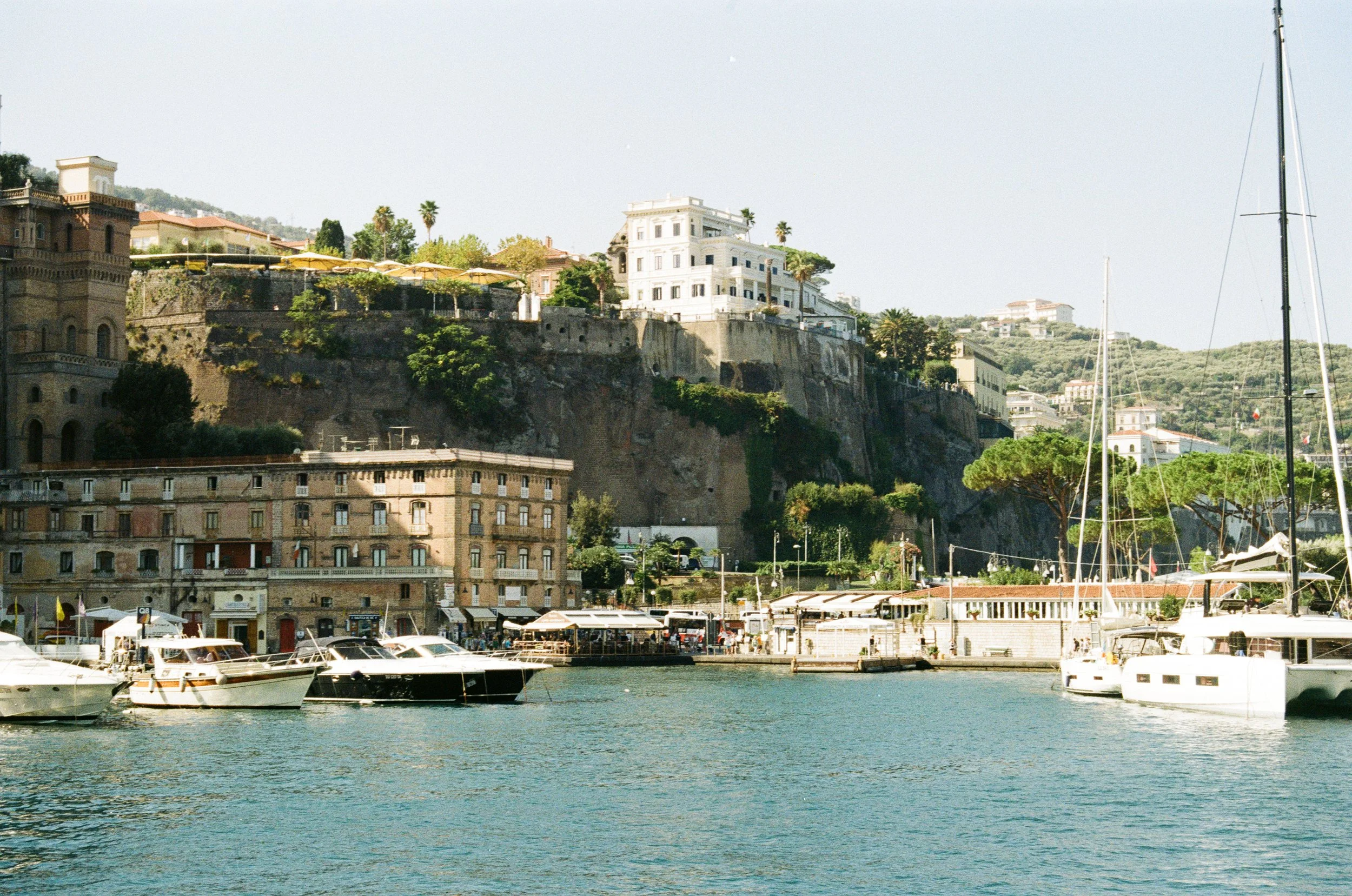 Photo of a marina with several boats docked, with buildings and houses on a hillside in the background.