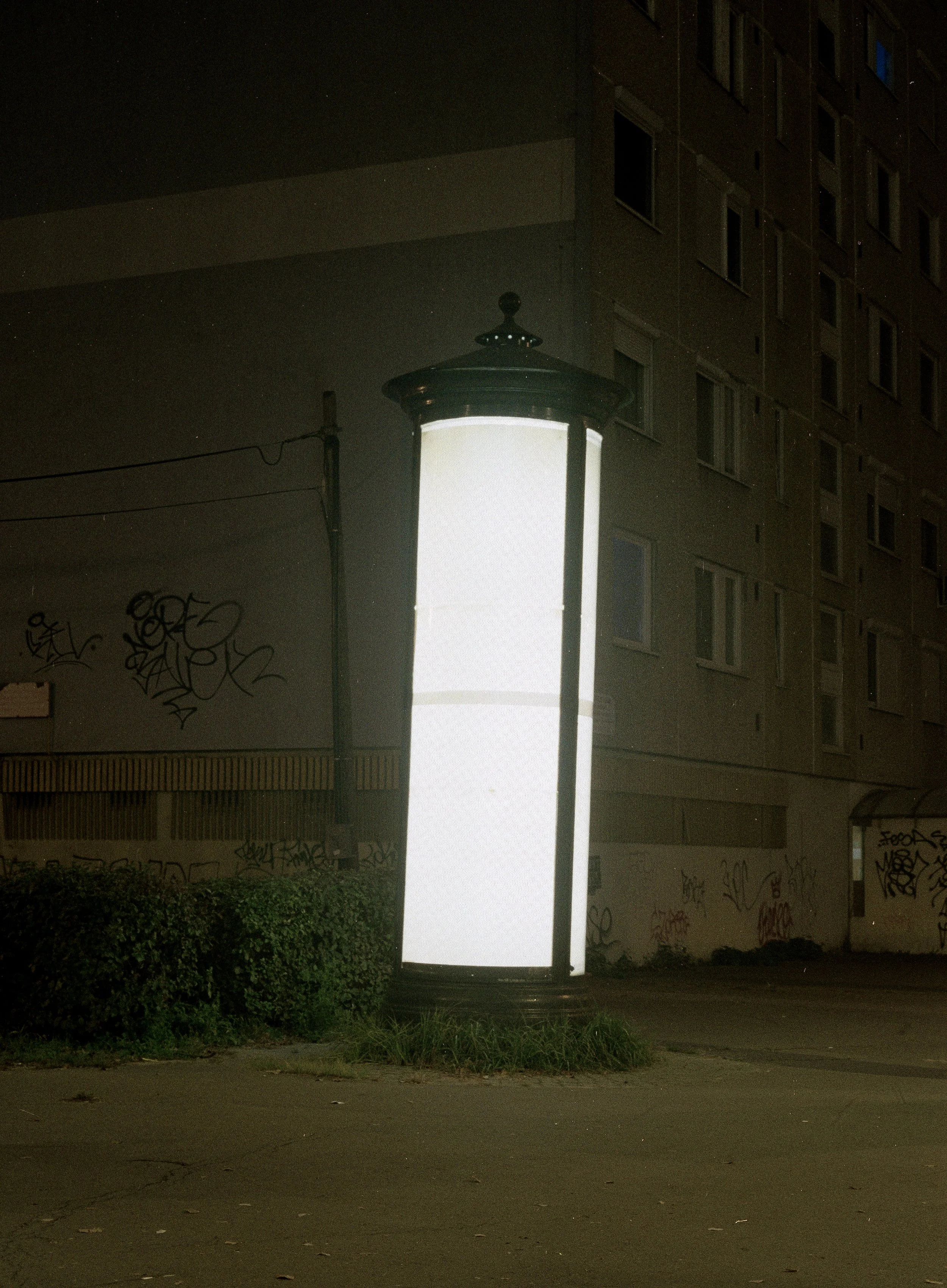 A tall, illuminated, white cylindrical street lamp on a sidewalk at night, with a building and graffiti in the background.