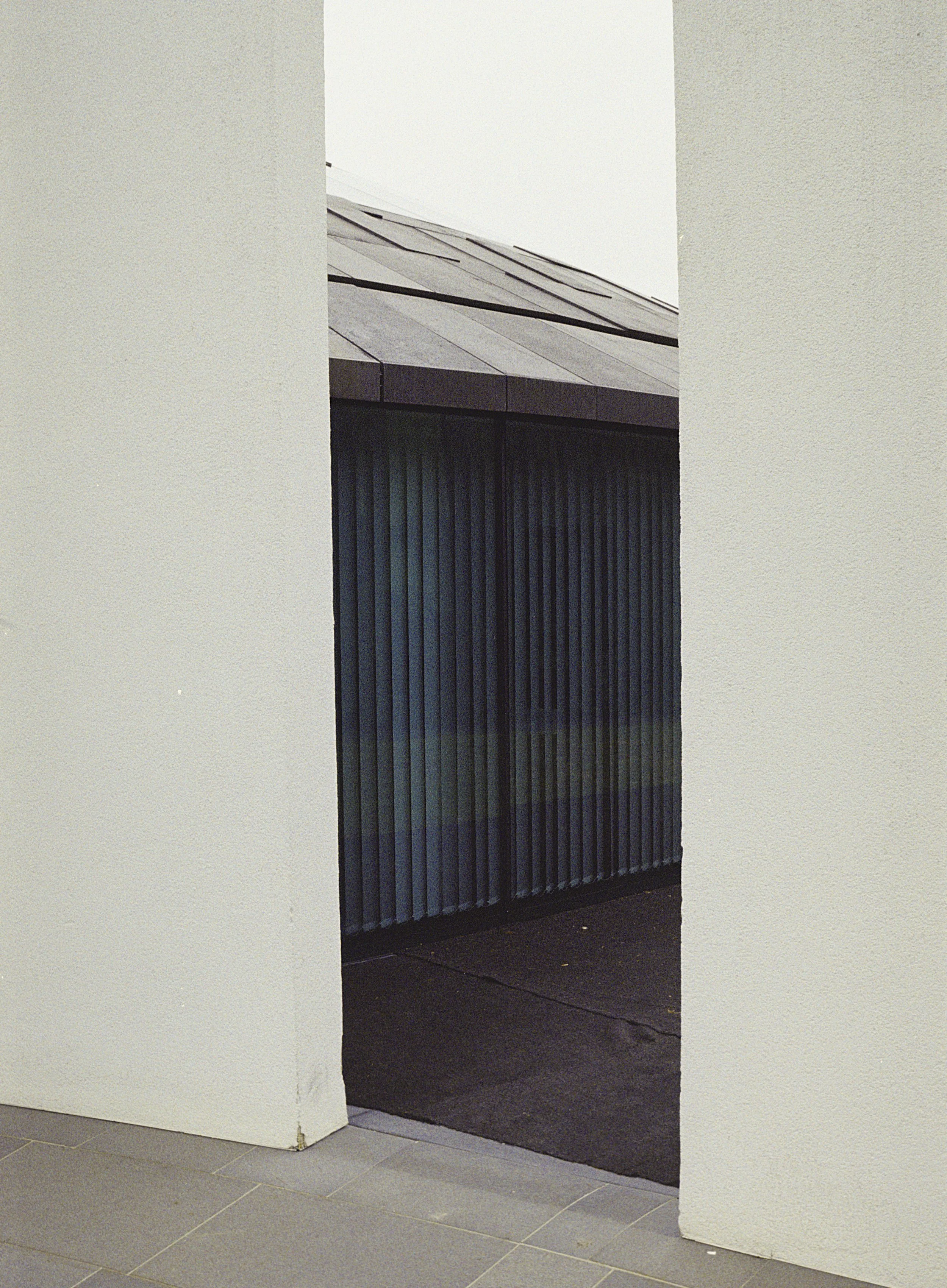 A corner of a building with a dark-colored slatted door or window and a white wall, with a tiled floor and cloudy sky visible above.