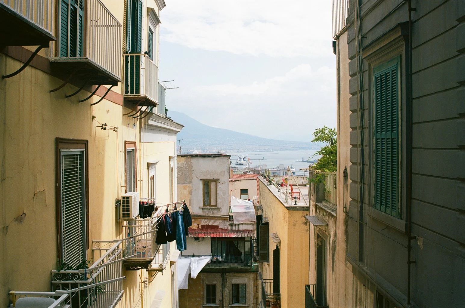 A view of narrow city streets with colorful buildings, laundry hanging on lines, and a distant body of water with ships and mountains.