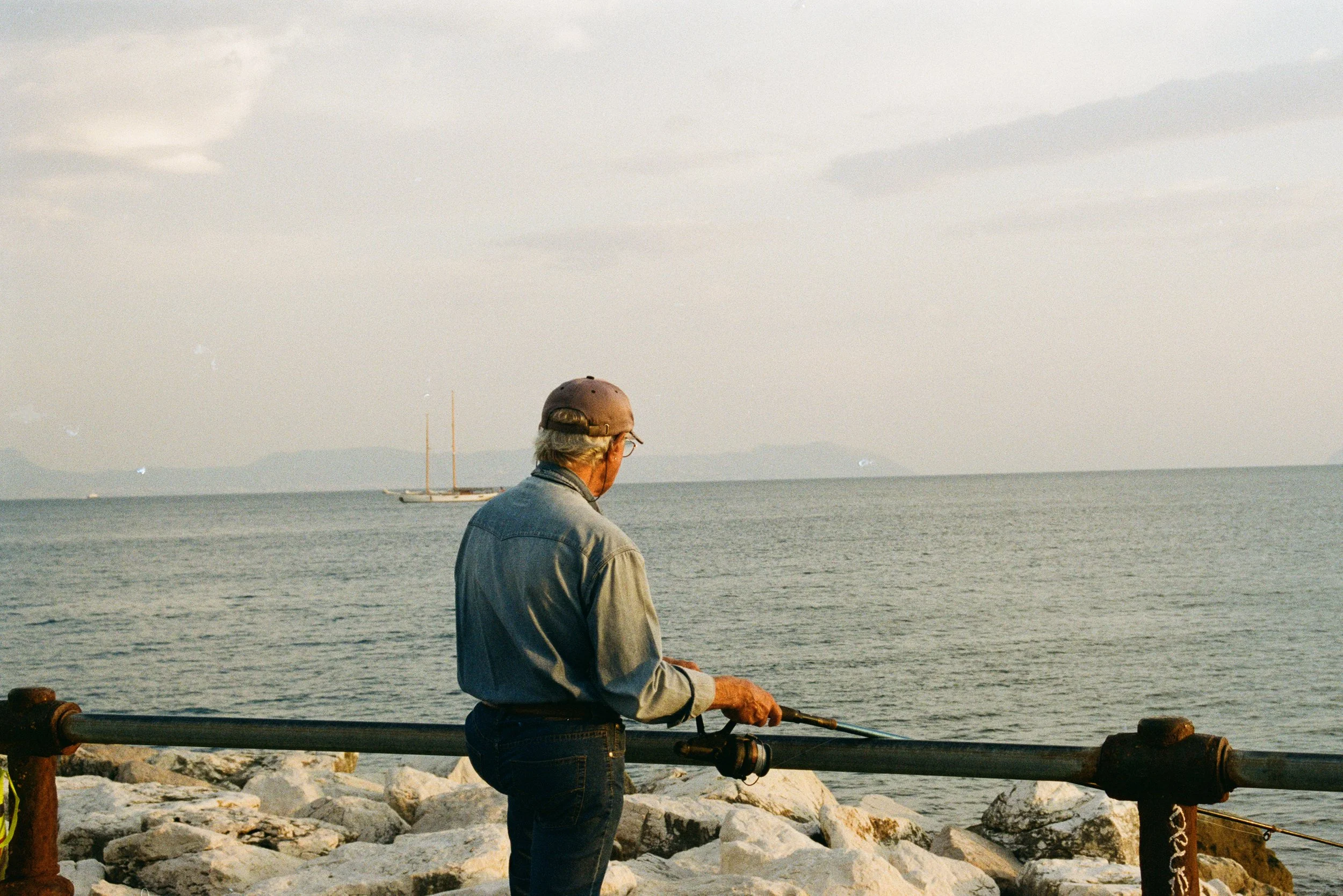 An elderly man with glasses, gray hair, wearing a cap and denim shirt, fishing off a rocky shoreline with a calm sea and sailboat in the distance.