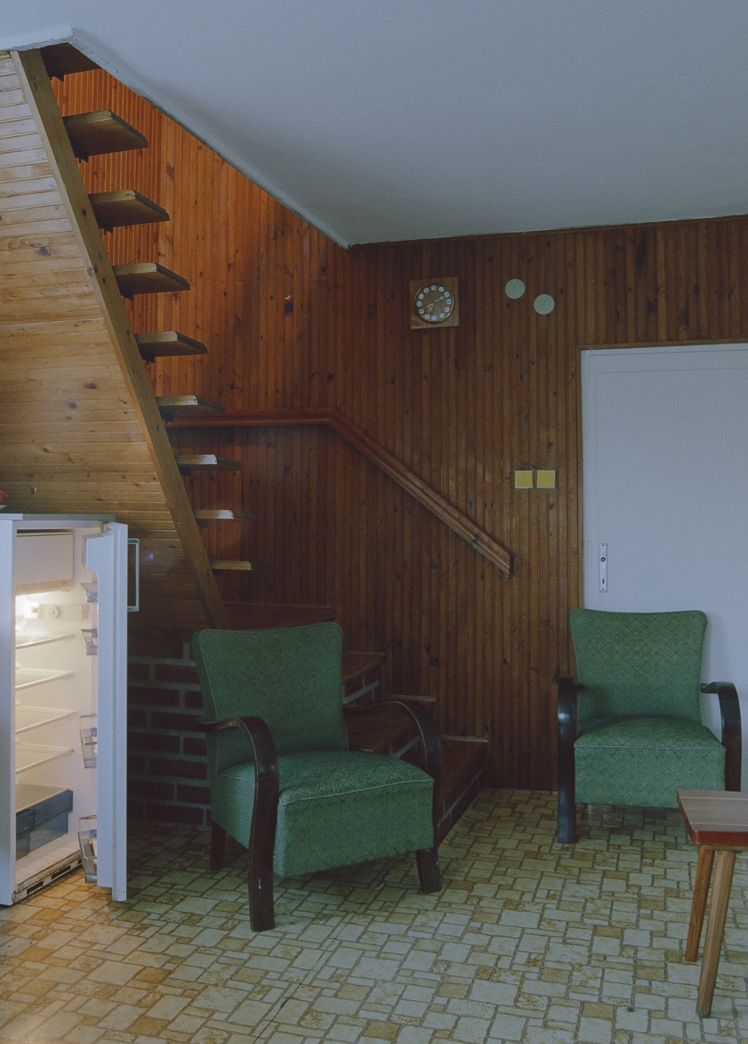 Interior view of a room with wooden panel walls, two green upholstered armchairs, a wooden staircase, and a small open refrigerator on the left. A clock is on the wall, and there is a white door on the right.