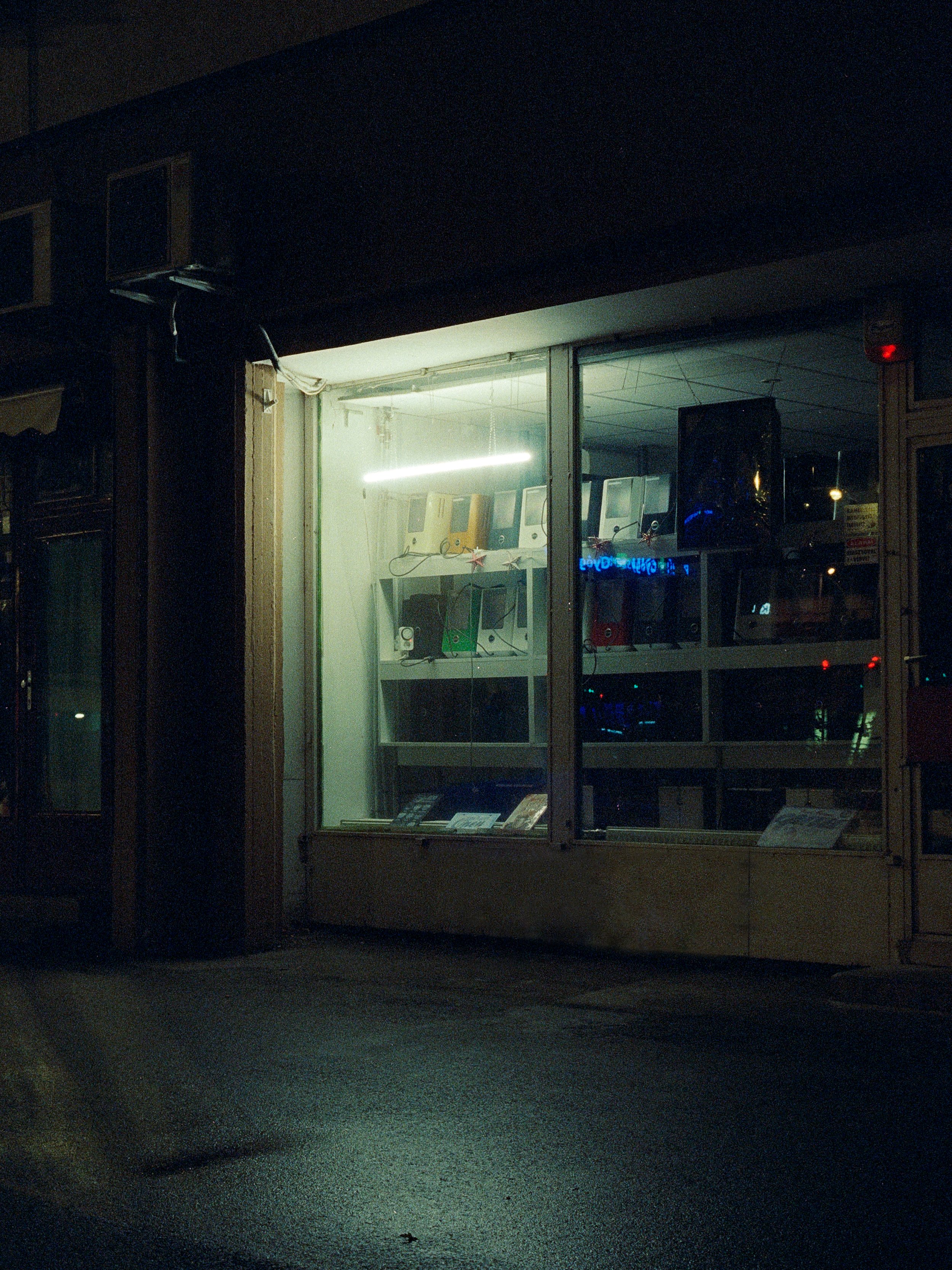 A storefront at night with a glass window displaying various electronic devices, including telephones and possibly portable radios, illuminated by a fluorescent light.