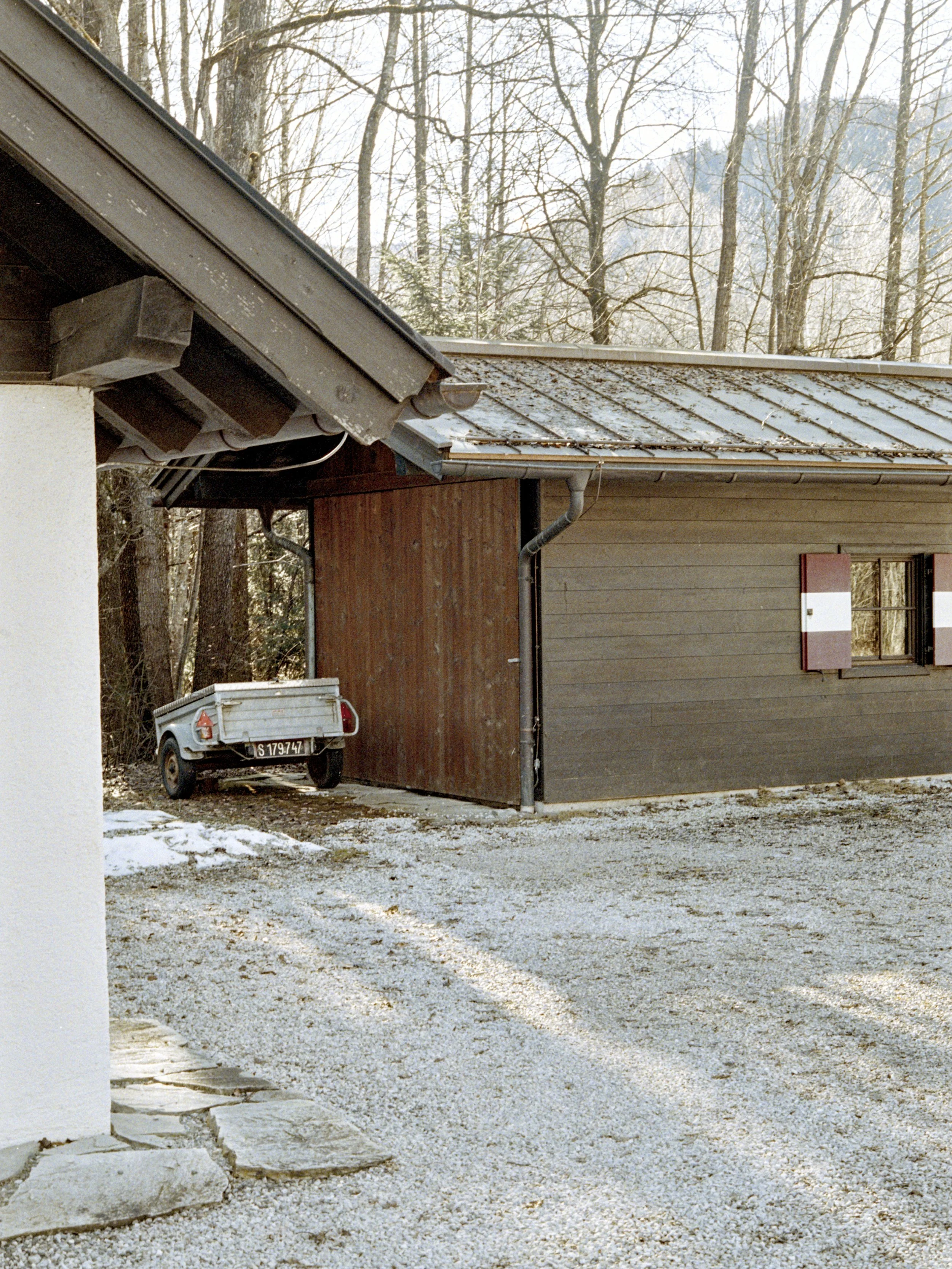 A gravel driveway in front of a house with wooden and stucco walls, a snow-dusted roof, and leafless trees in the background.