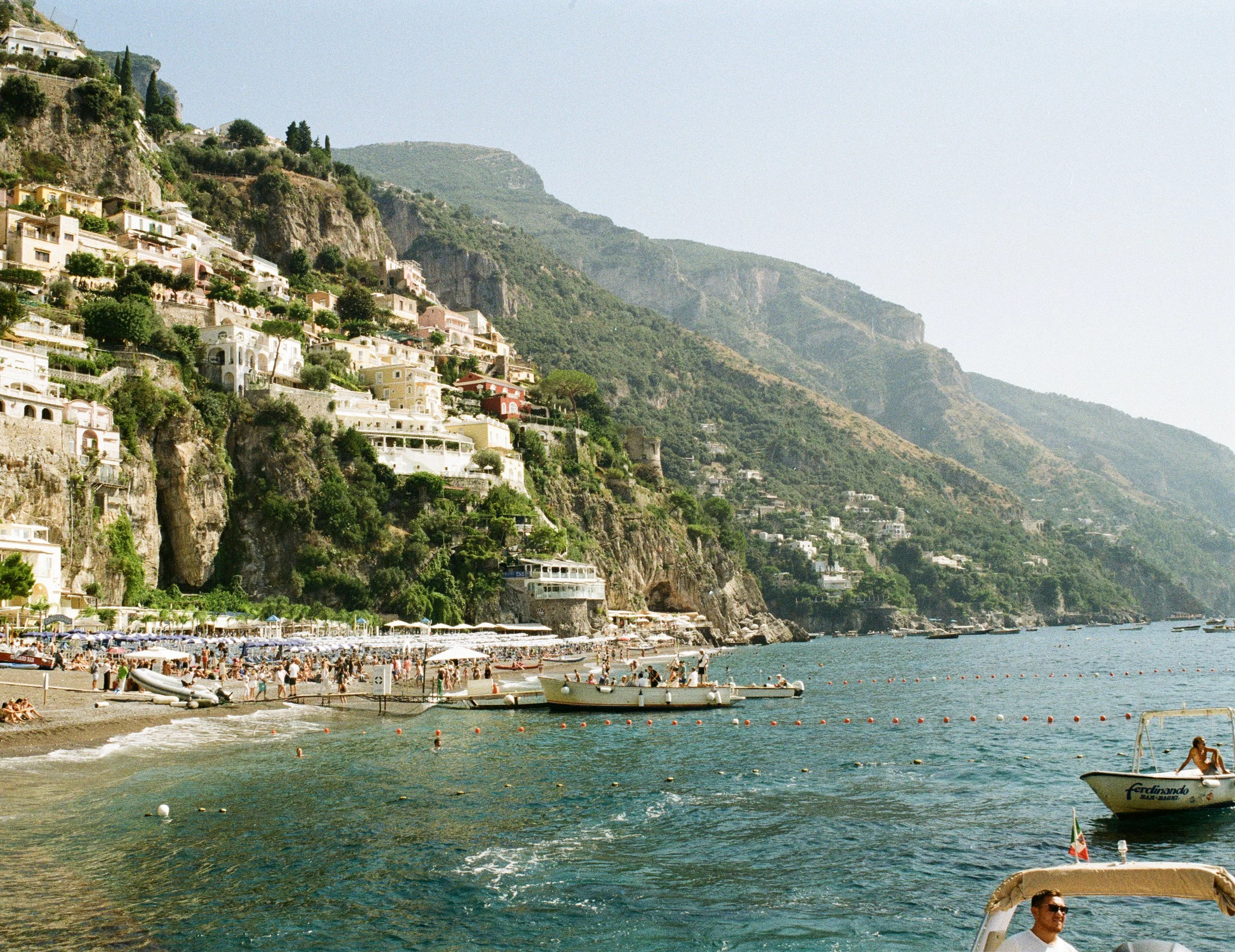 Coastal scene of a beach with umbrellas and boats, and a hillside with colorful residential buildings in the background.