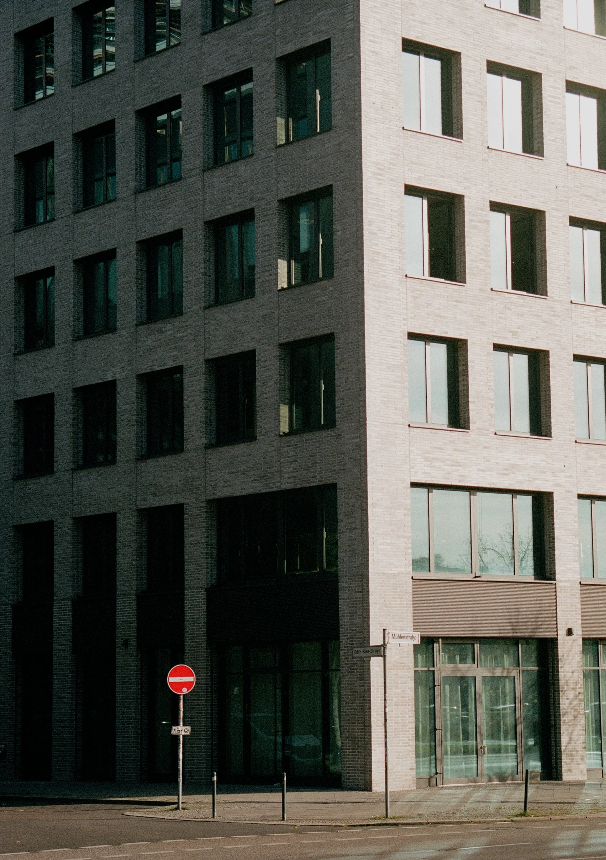 A modern multi-story building with a facade of gray bricks and large rectangular windows, located on a city street with a no entry traffic sign and street signs.