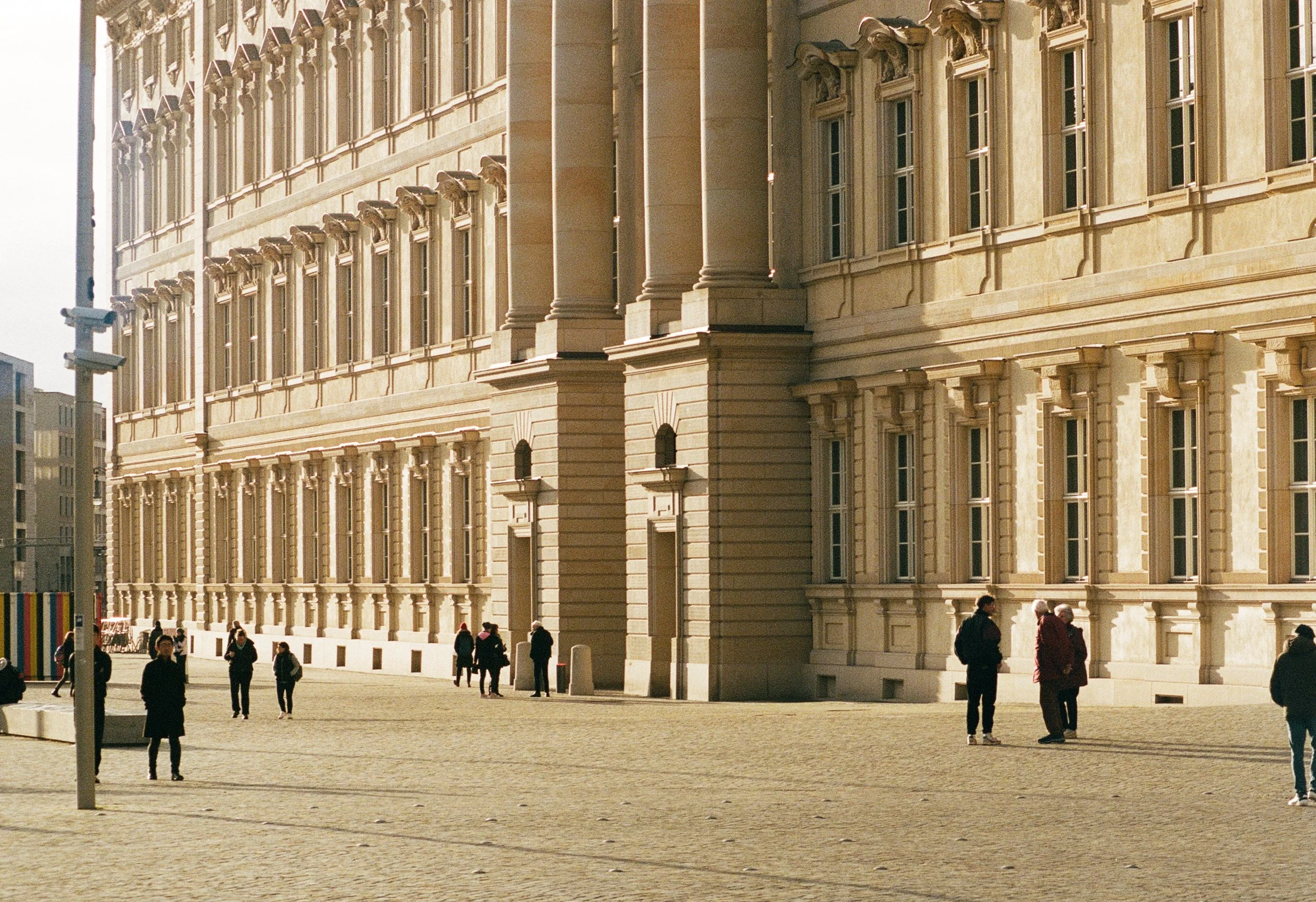A large historic building with classical architecture and detailed stone work, with several people walking and standing outside on a sunny day.