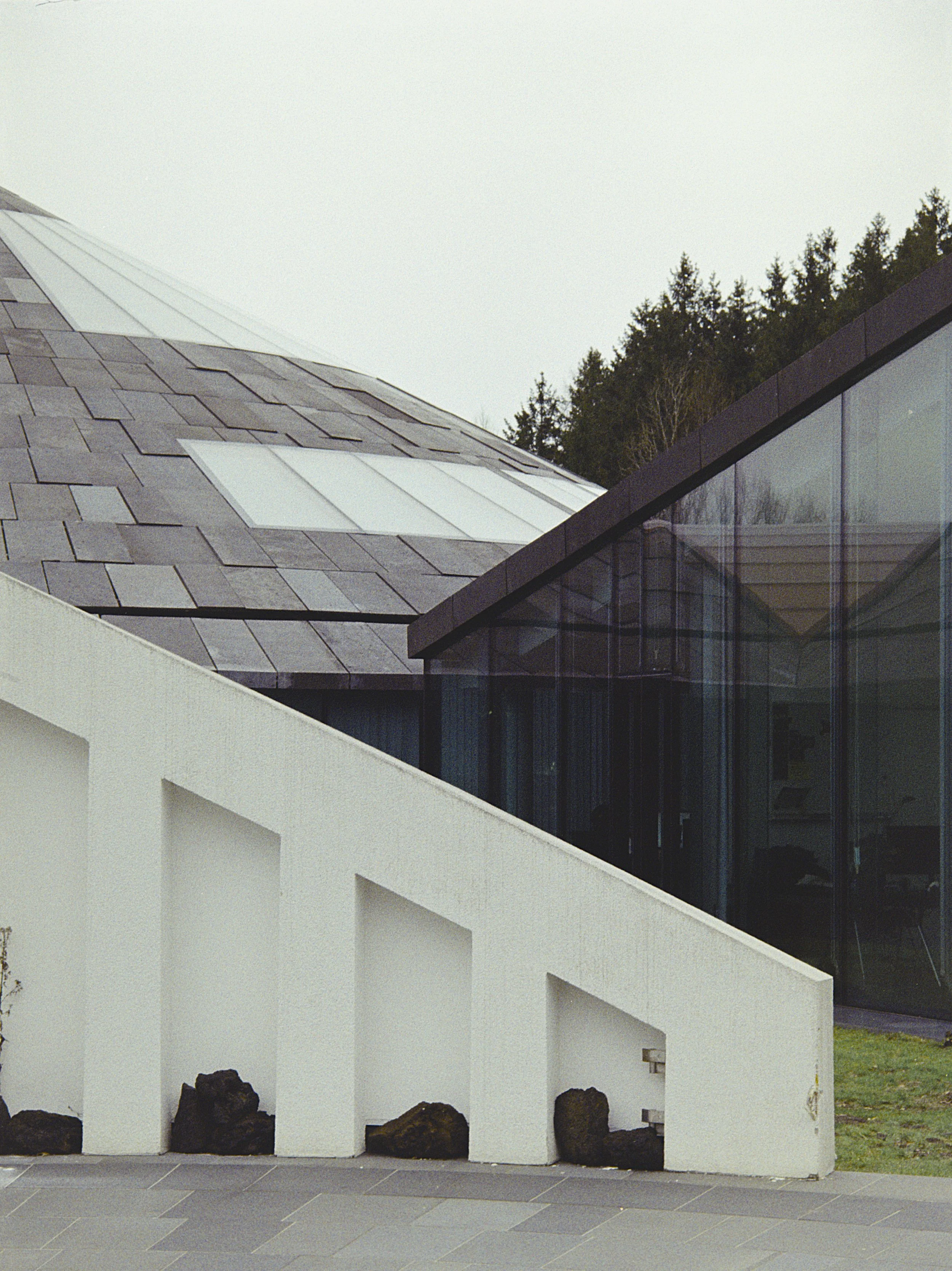 Modern architectural building with a white concrete wall, large rocks at the base, and a roof covered in gray slate tiles with skylights. A glass section of the building reflects the sky and trees.