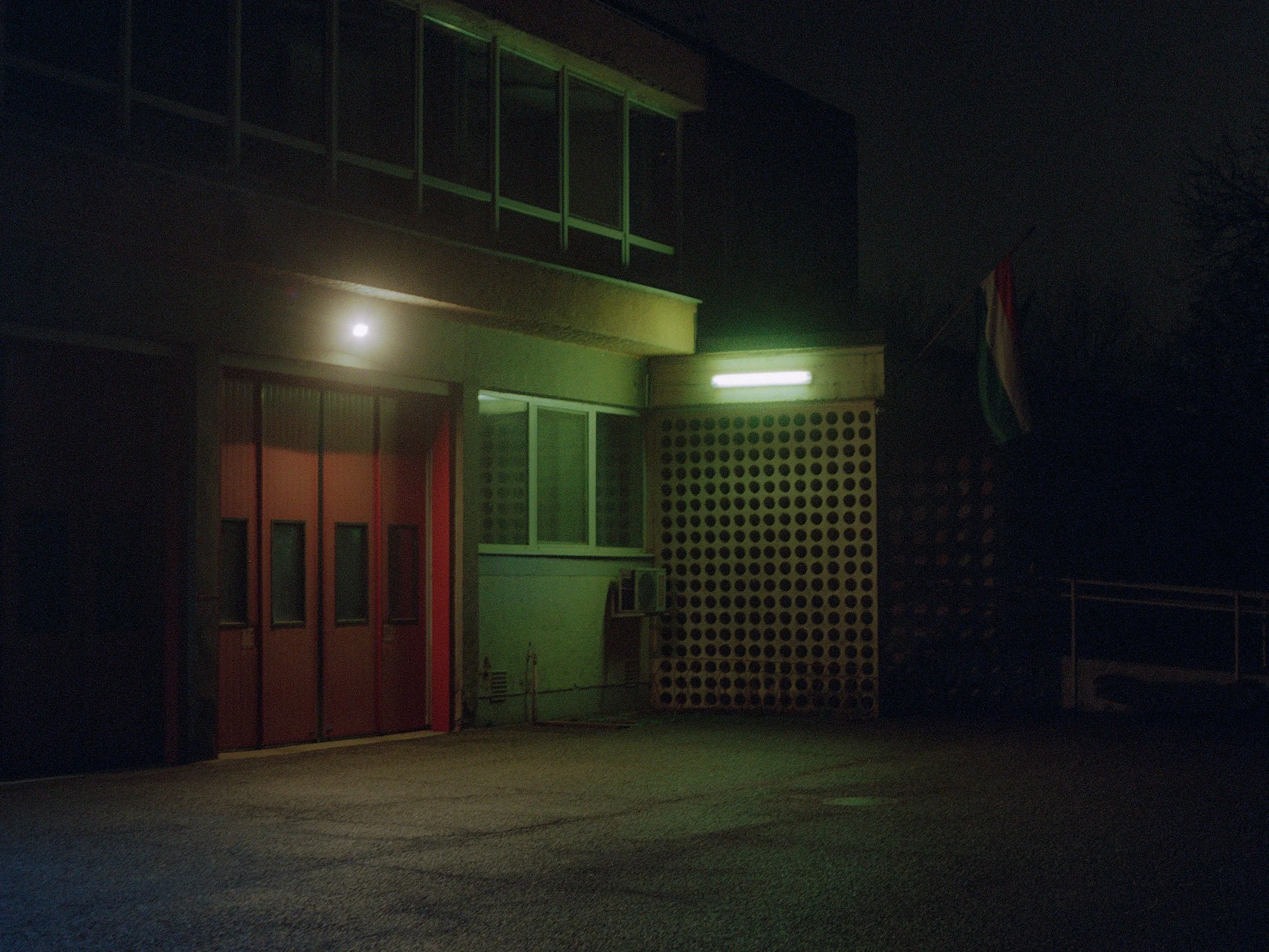 A dark night scene of a building with red double doors, illuminated by green and white lights, with a flag on the right side of the building.