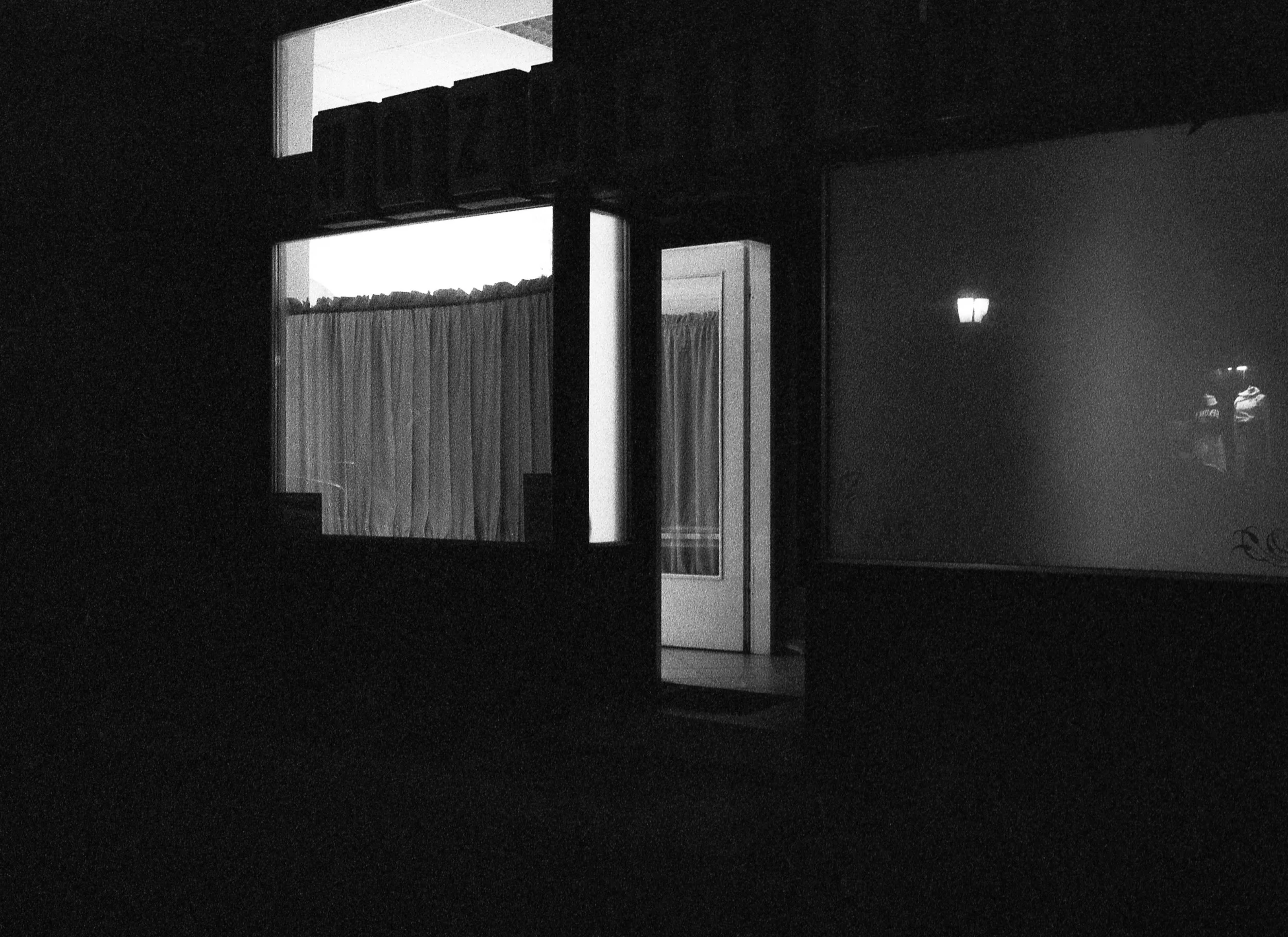 Dark storefront with a bright doorway and two windows, one with closed curtains and another reflecting streetlights.
