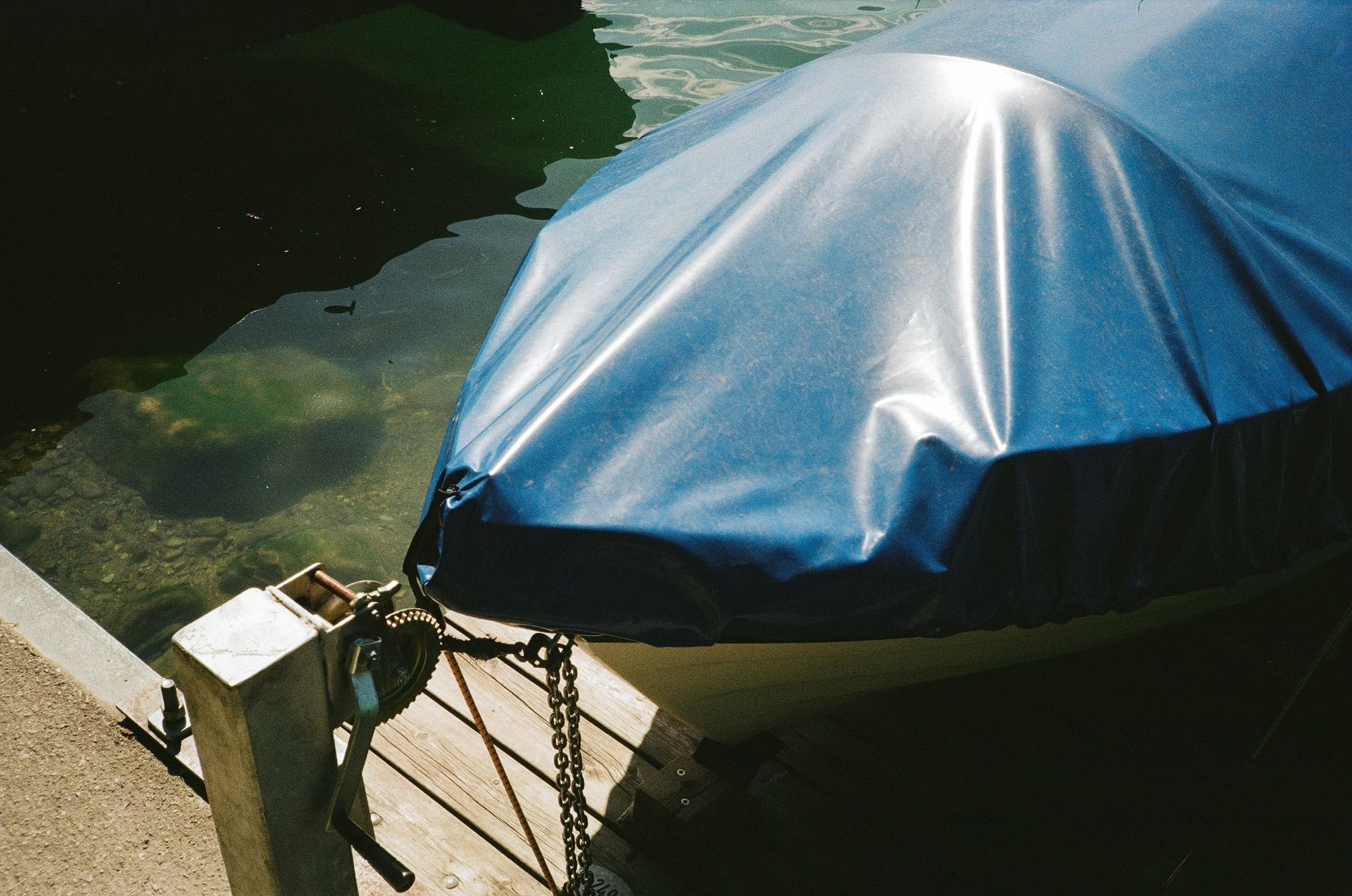 A boat covered with a blue tarp is tied to a wooden dock with a chain, floating on calm water.