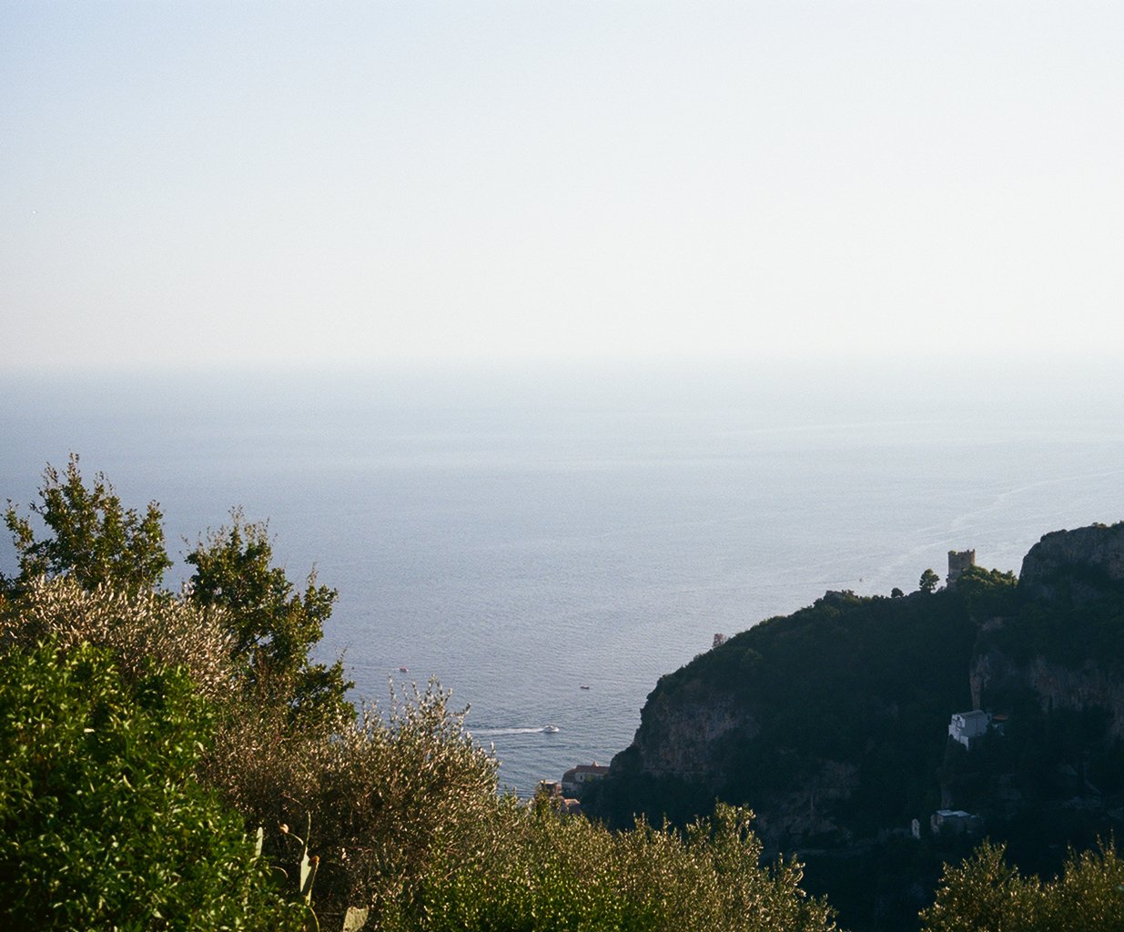 A scenic view of the sea with a boat sailing, surrounded by green trees and a rocky hillside with a castle on top.