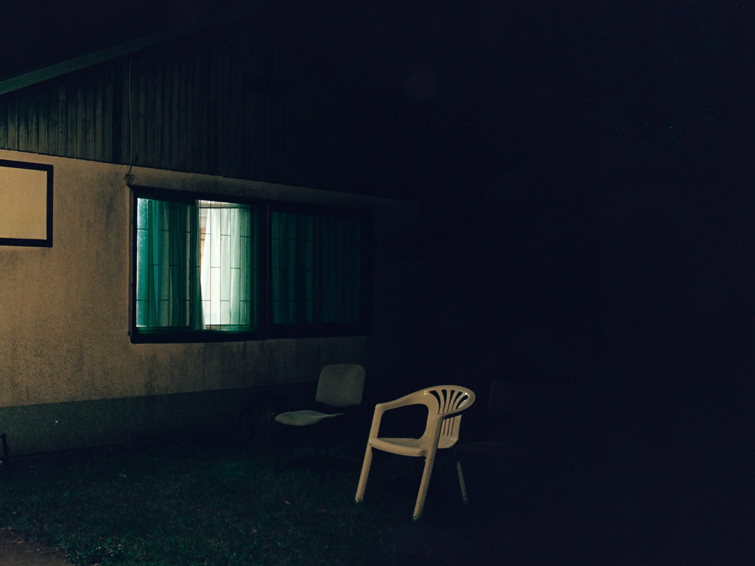 Nighttime view of a house exterior with a window and two lawn chairs outside, illuminated slightly by indoor light.
