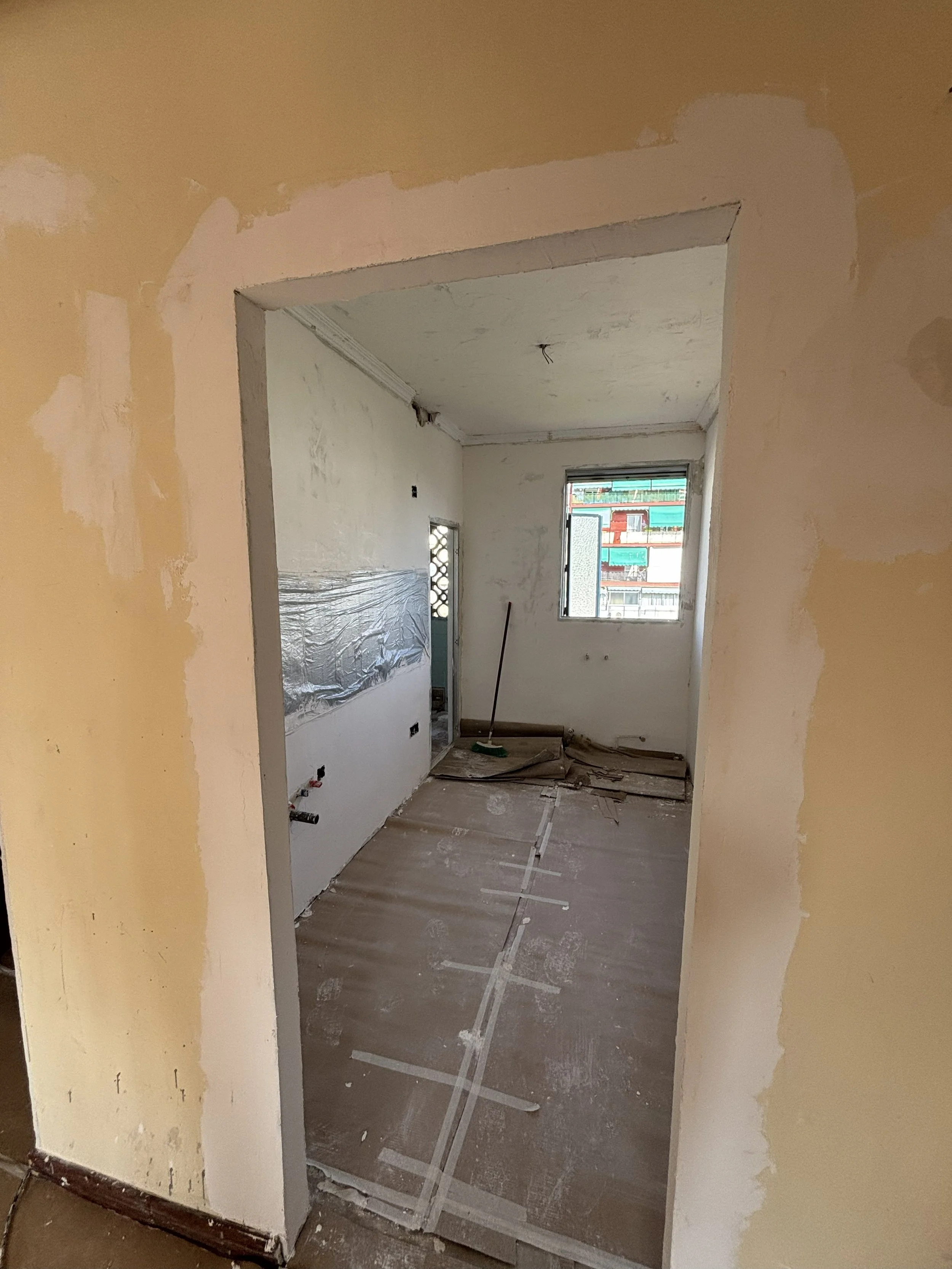 Interiores de una cocina en construcción con paredes sin pintar, piso cubierto con cartón para protección, y una ventana que da a un edificio cercano.