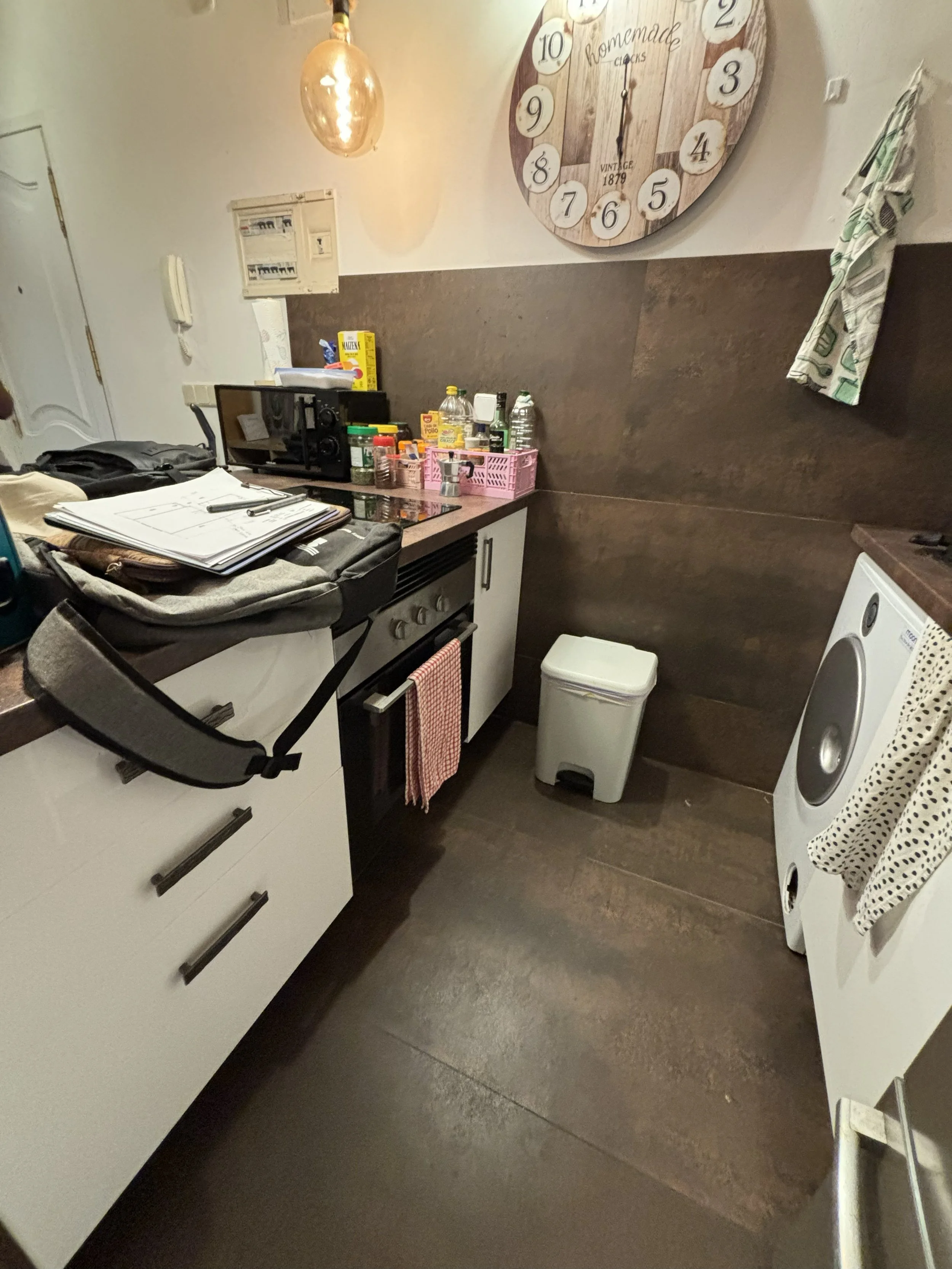 Cocina pequeña con gabinetes blancos, utensilios y alimentos en la encimera, una lavadora, un bote de basura, una pared con reloj grande de madera y una toalla colgada, y un foco colgante