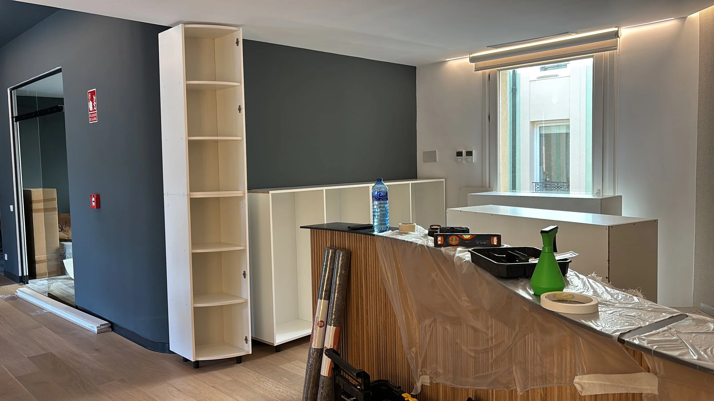 Interior en remodelación con estantes blancos, mesa cubierto con plástico, botellas de agua, herramienta y equipo de construcción, pared gris y ventana.