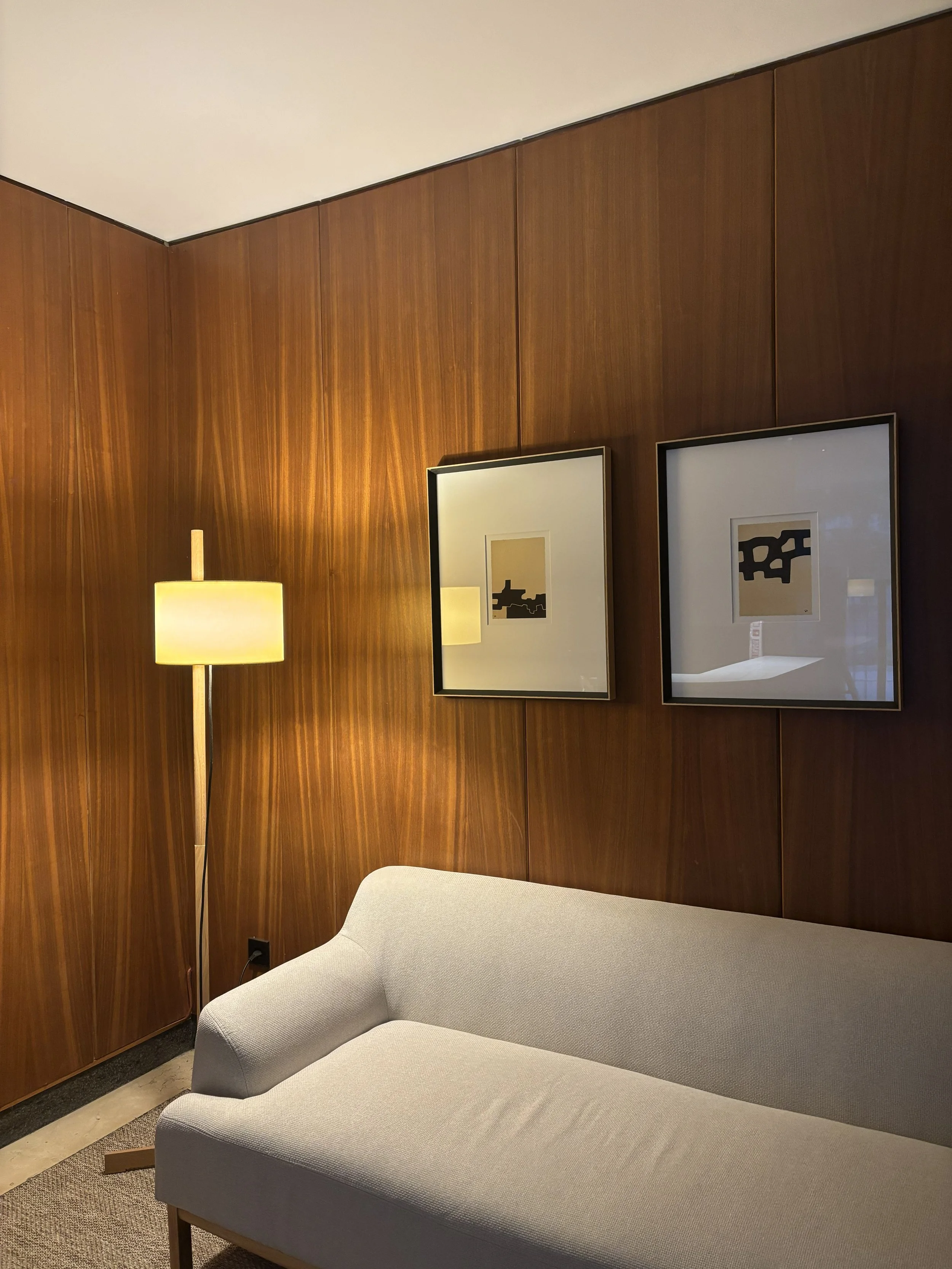 Sala con pared de madera, un sofá gris claro y una lámpara de pie con pantalla beige, dos cuadros contemporáneos en la pared.