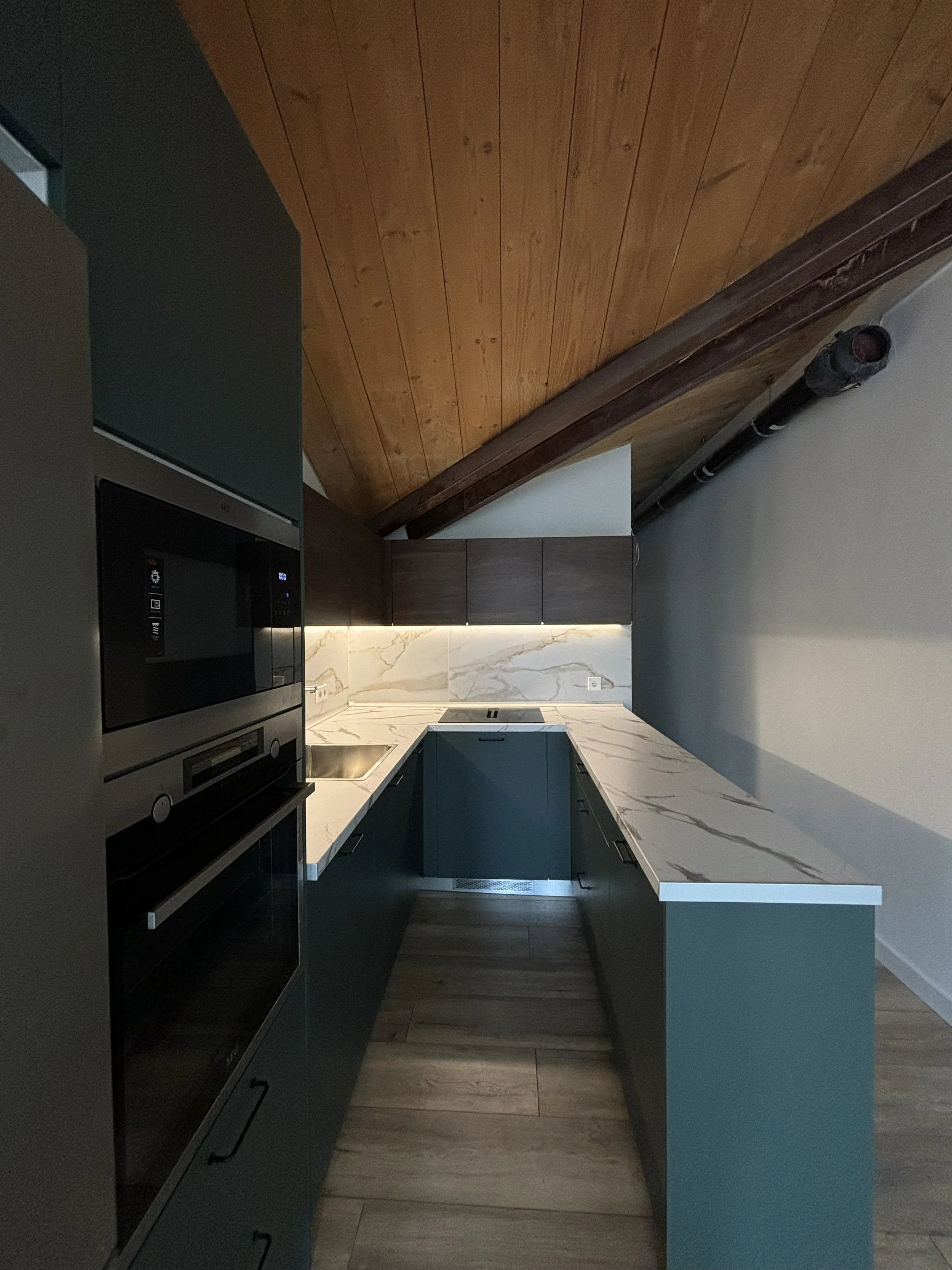 Cocina moderna con gabinetes en color gris oscuro, encimera de mármol blanco con vetas grises, paredes blancas, techo de madera y tuberías expuestas.