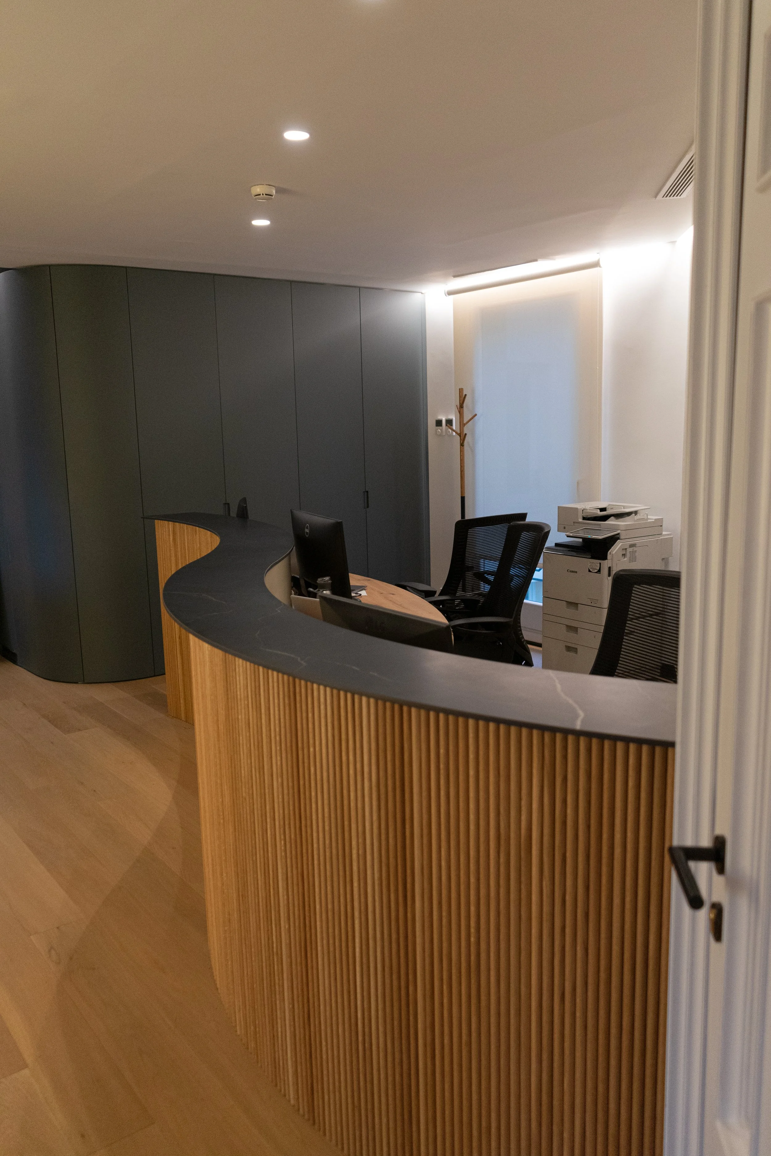 Mostrador de recepción de oficina con computadora, sillas de oficina y una planta de esquina, con paredes blancas y puertas de madera.