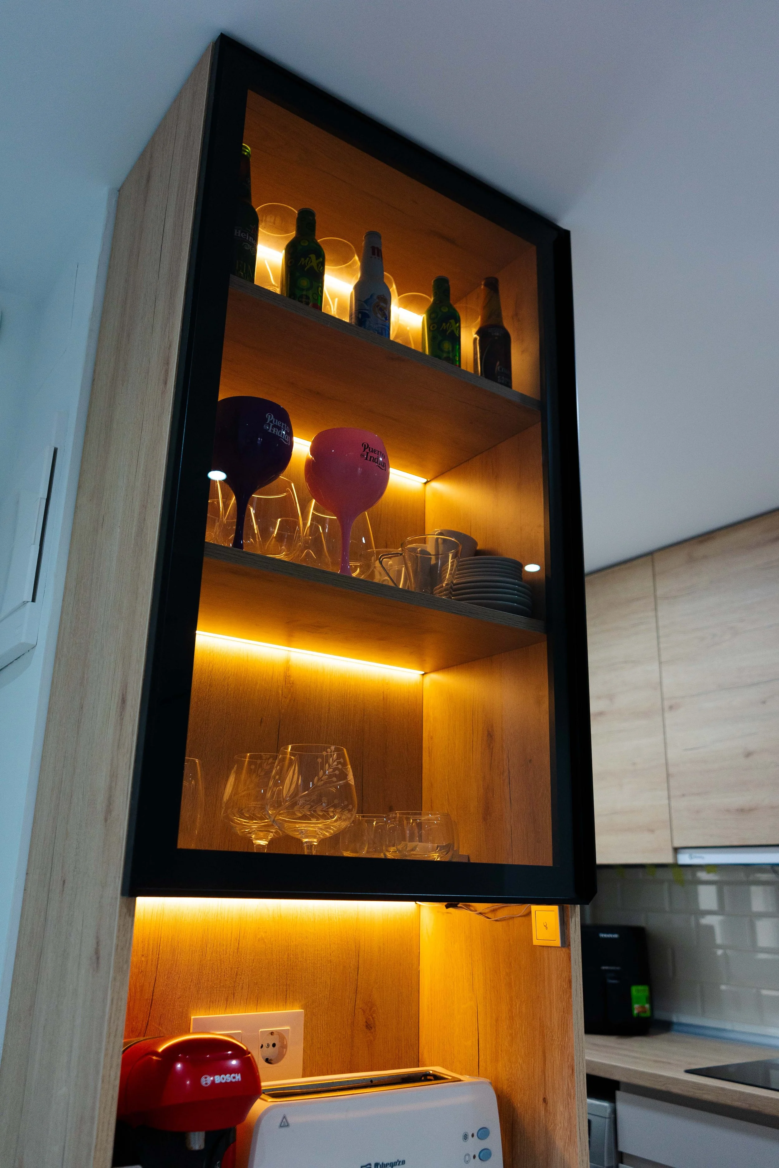 Estantes con botellas, tazas y copas de vino en una cocina moderna con iluminación cálida.